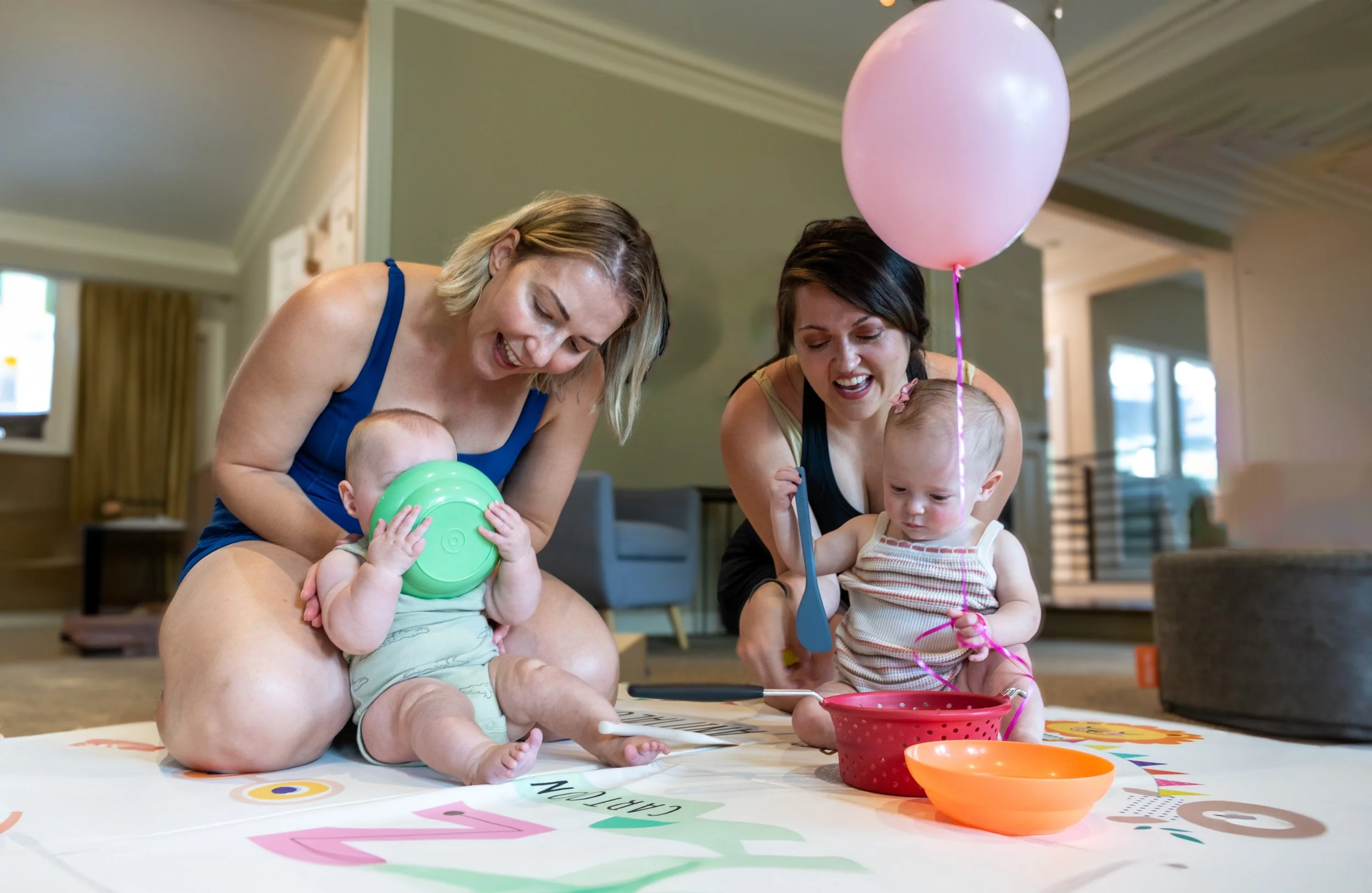 Two women and two young children sitting on a floor playing a game with colorful bowls and small objects, with balloons nearby in an indoor space at a Tiny Explorers playgroup