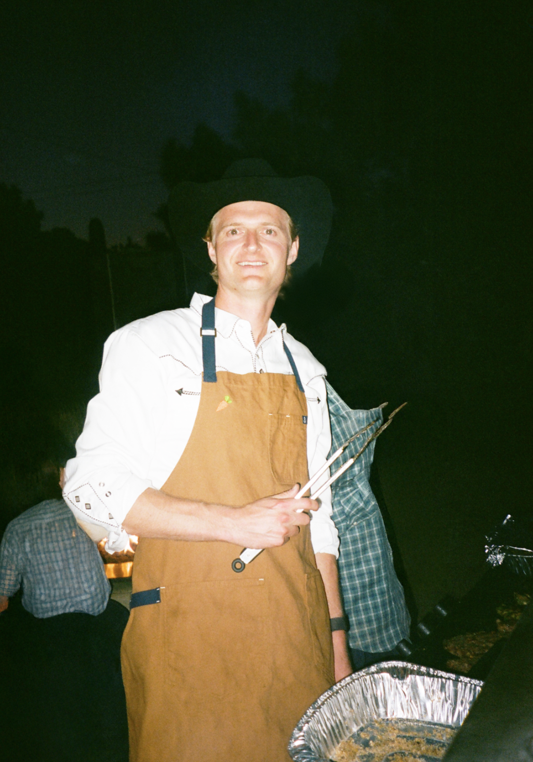 Man wearing a cowboy hat and white shirt with rolled-up sleeves, cooking with tongs at an outdoor event during nighttime.