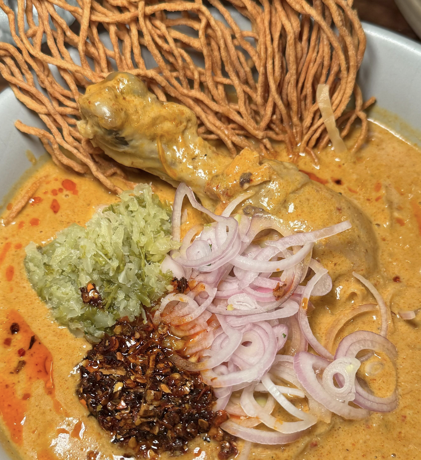 Bowl of spicy ramen with chicken drumstick, fried noodles, chopped pickled vegetables, sliced onions, chopped garlic, and chili paste in broth.