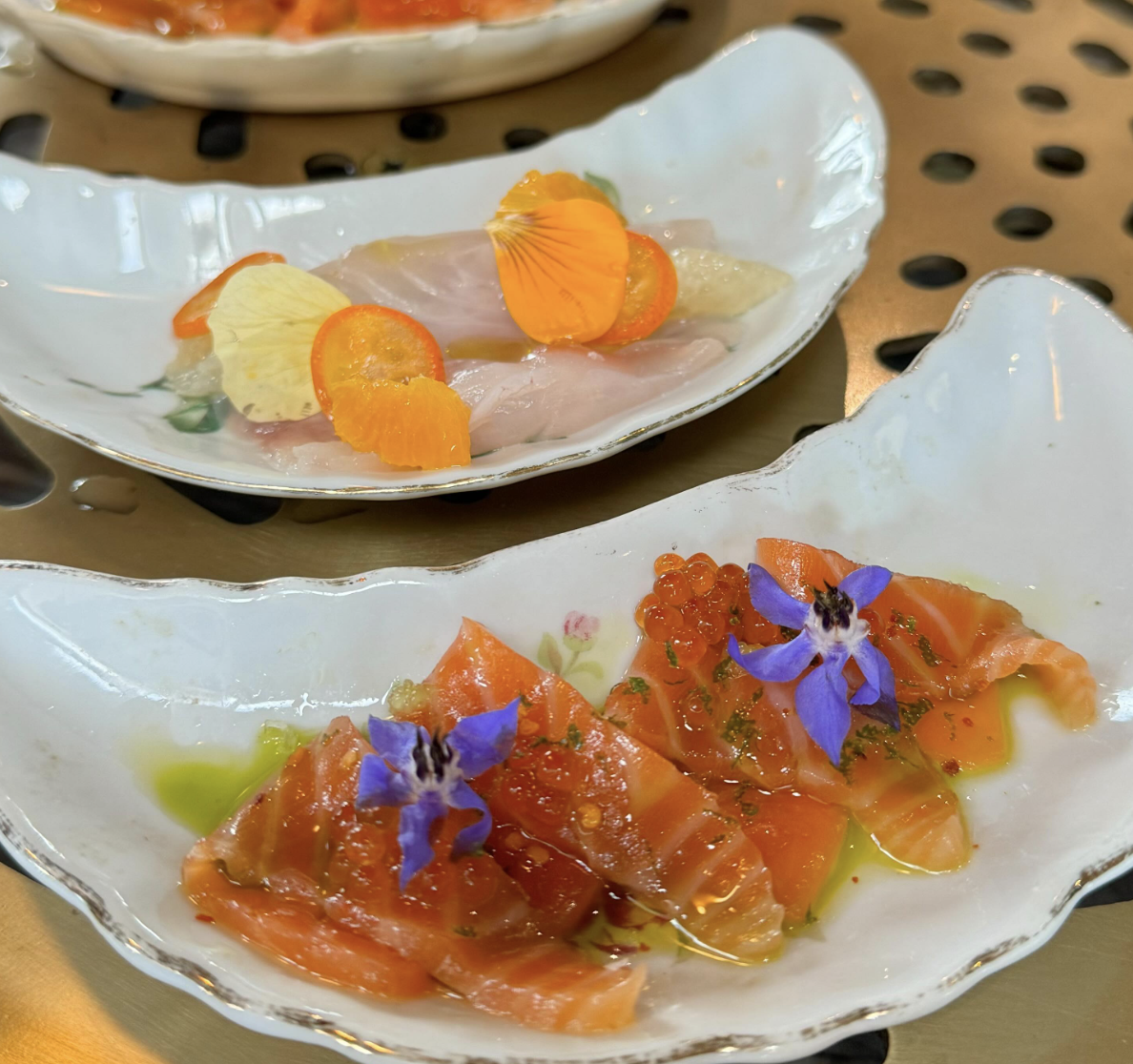 Two white ceramic plates with smoked salmon topped with edible flowers and herbs. The plate in the foreground has orange and purple flowers with a drizzle of green oil, while the plate in the background has slices of fish garnished with orange and yellow edible flower petals.