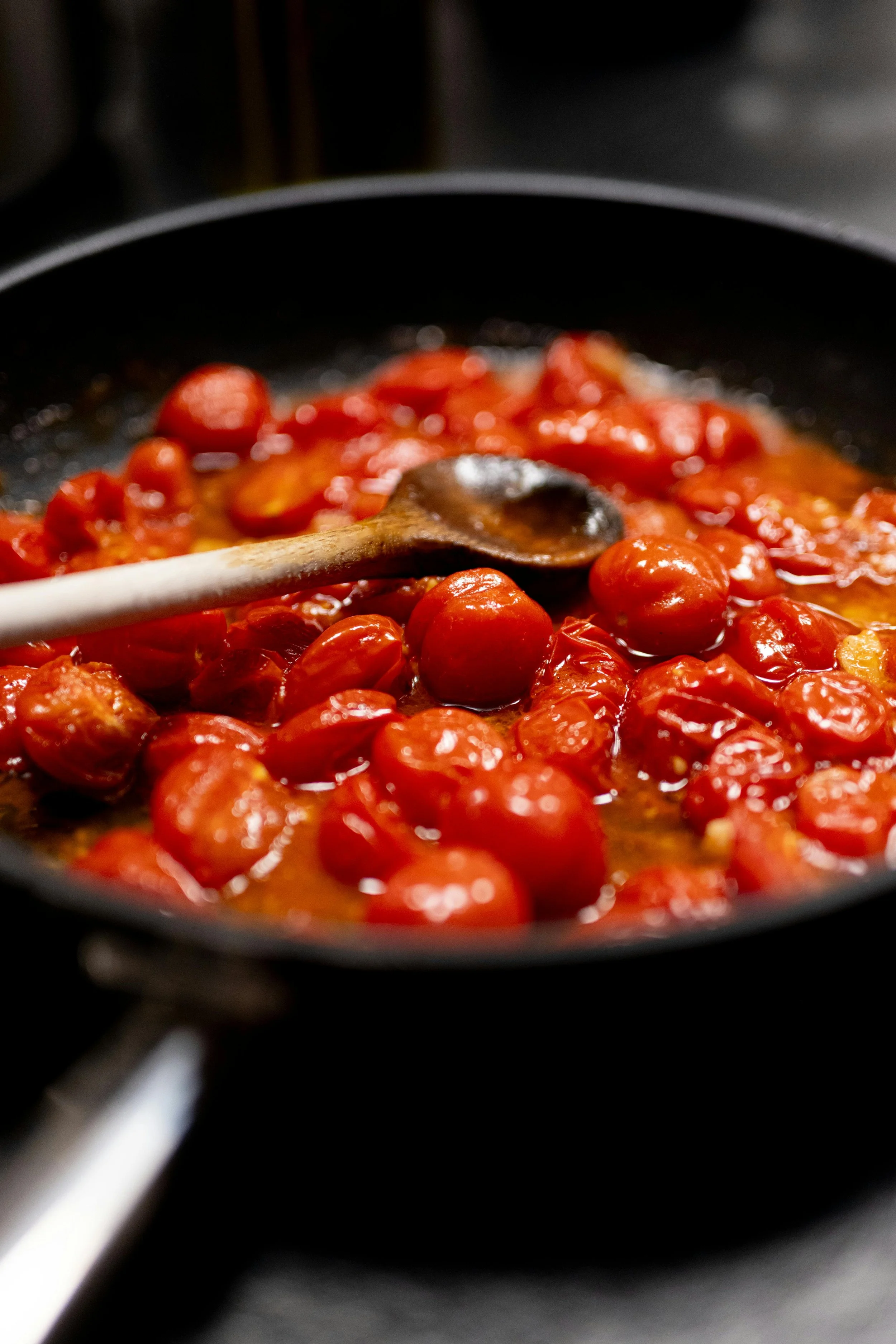 Cherry tomatoes cooking in a skillet on a stove with a wooden spoon stirring.