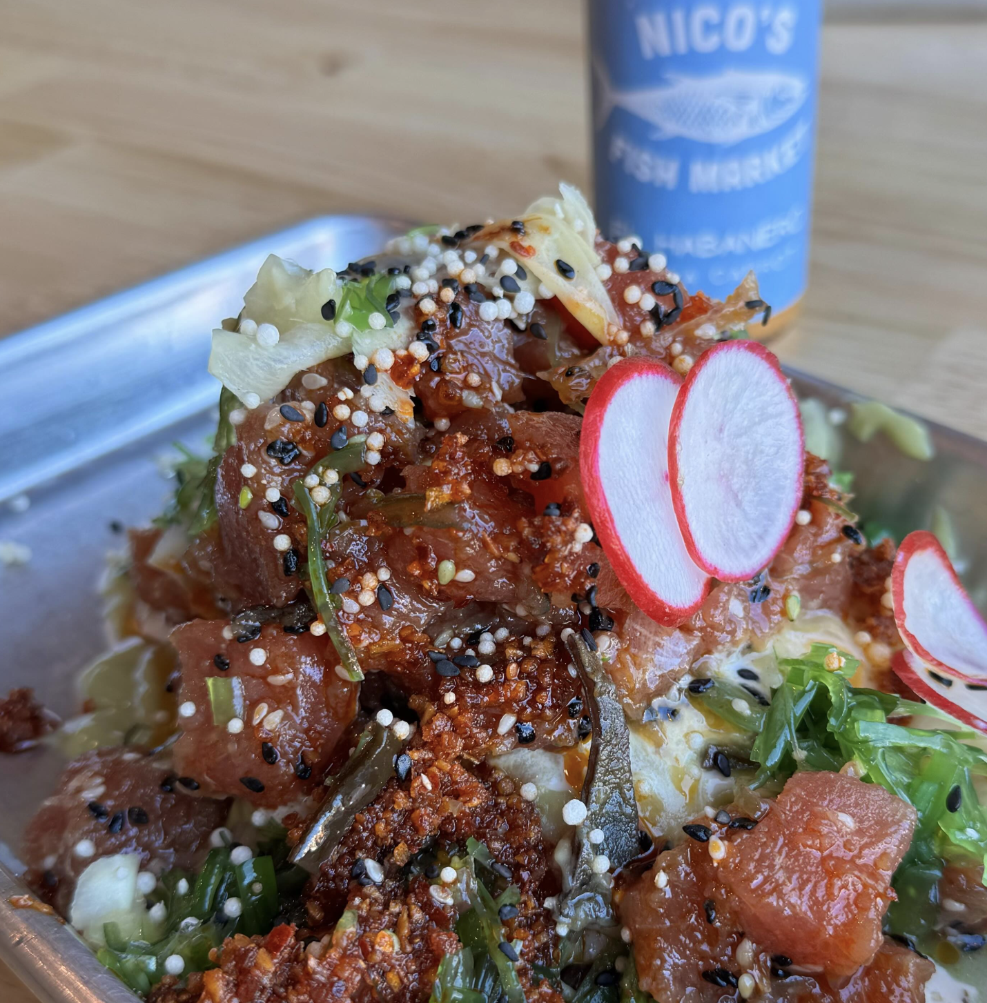Close-up of chopped raw fish with sliced radishes, seaweed, and sesame seeds on a metal tray, with a blue can in the background.