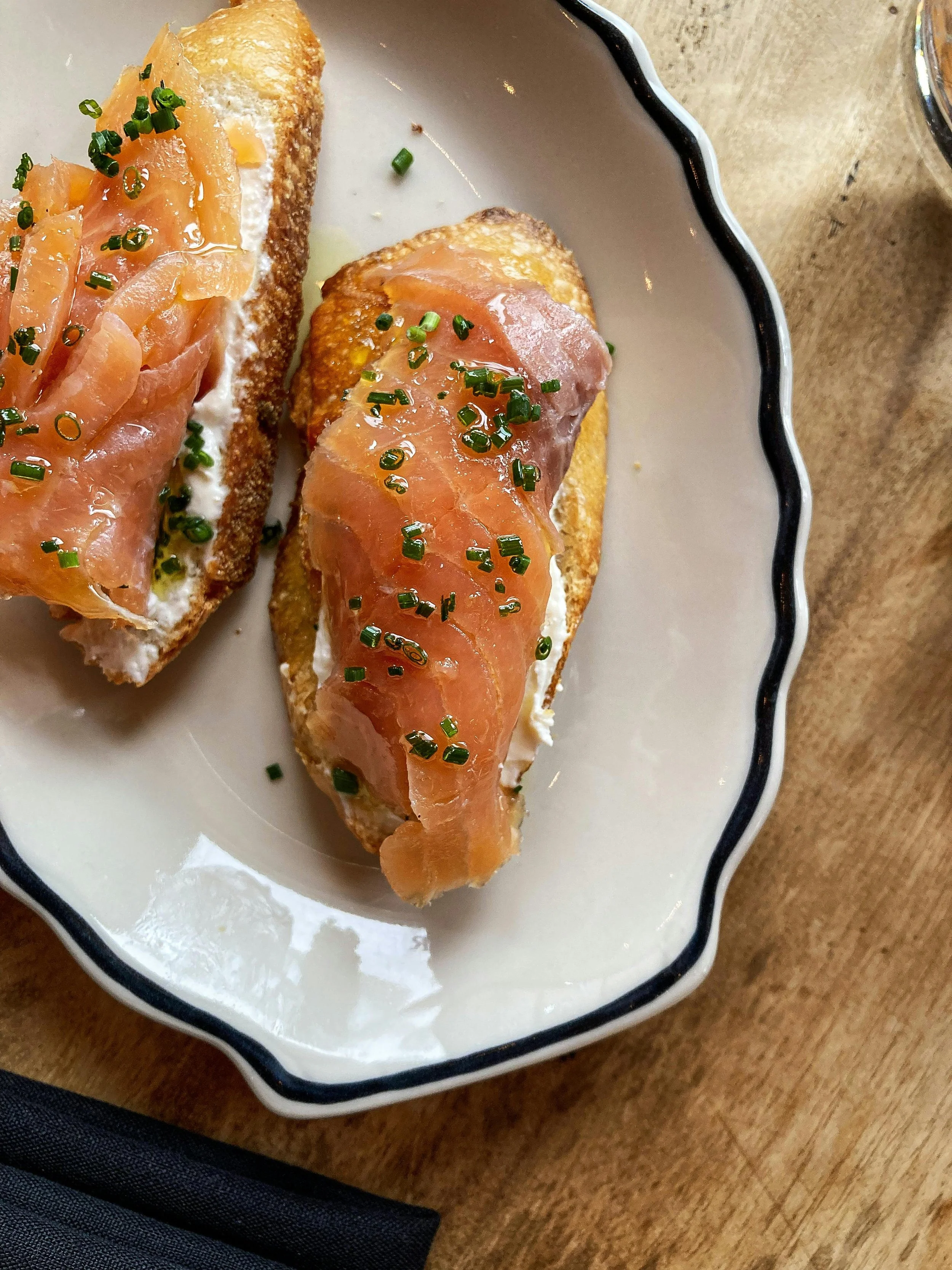Two pieces of crostini topped with smoked salmon, cream cheese, and chopped chives on a white plate with black trim.