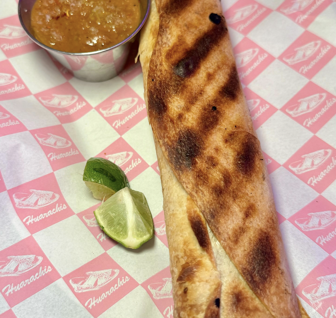 A Mexican burrito with grilled marks, served with lime wedges, salsa in a metal cup, on pink and white checkered paper.