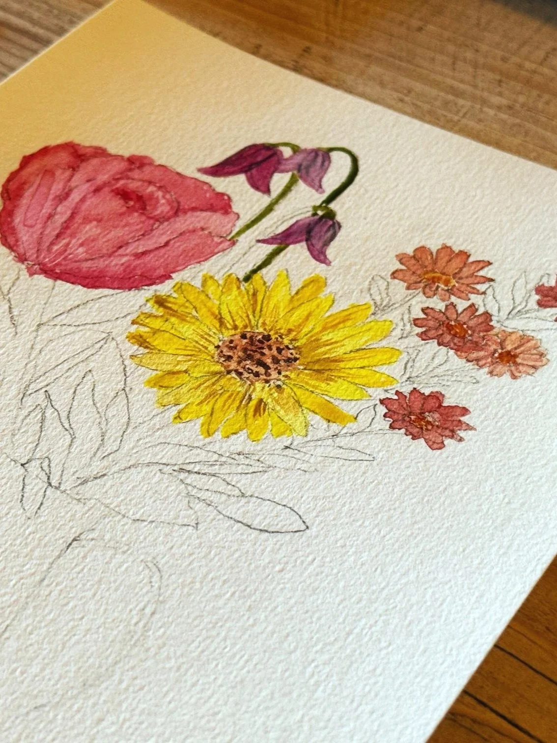 Partially completed watercolour painting of various flowers, including a pink rose, purple tulips, a yellow sunflower, and small orange daisies, on textured white paper.