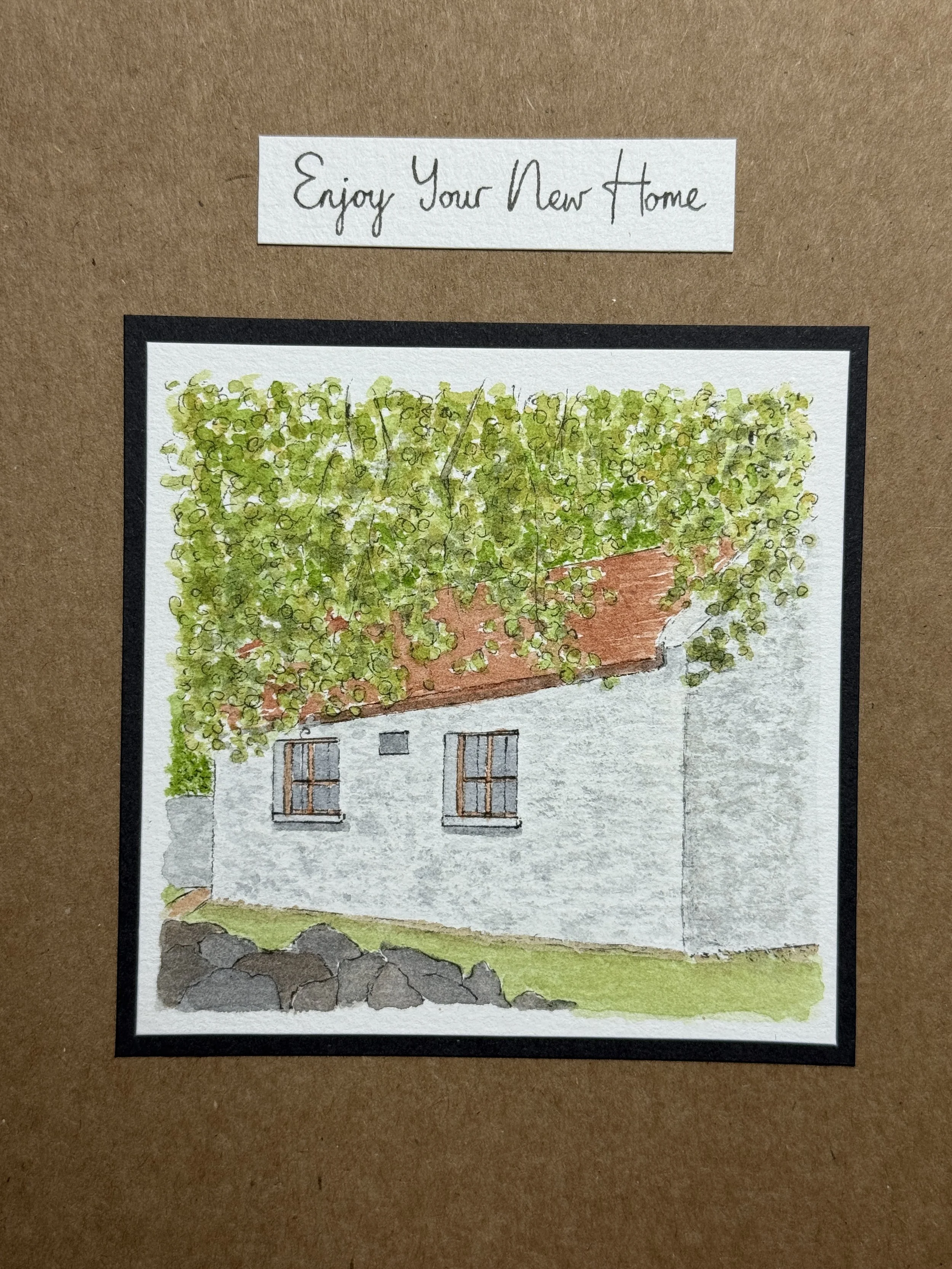 A watercolour painting of a small house with white walls, two windows with brown frames, and a red-tiled roof. Greenery covers part of the house and the ground is grassy with some rocks. A note above the painting says, "Enjoy Your New Home."