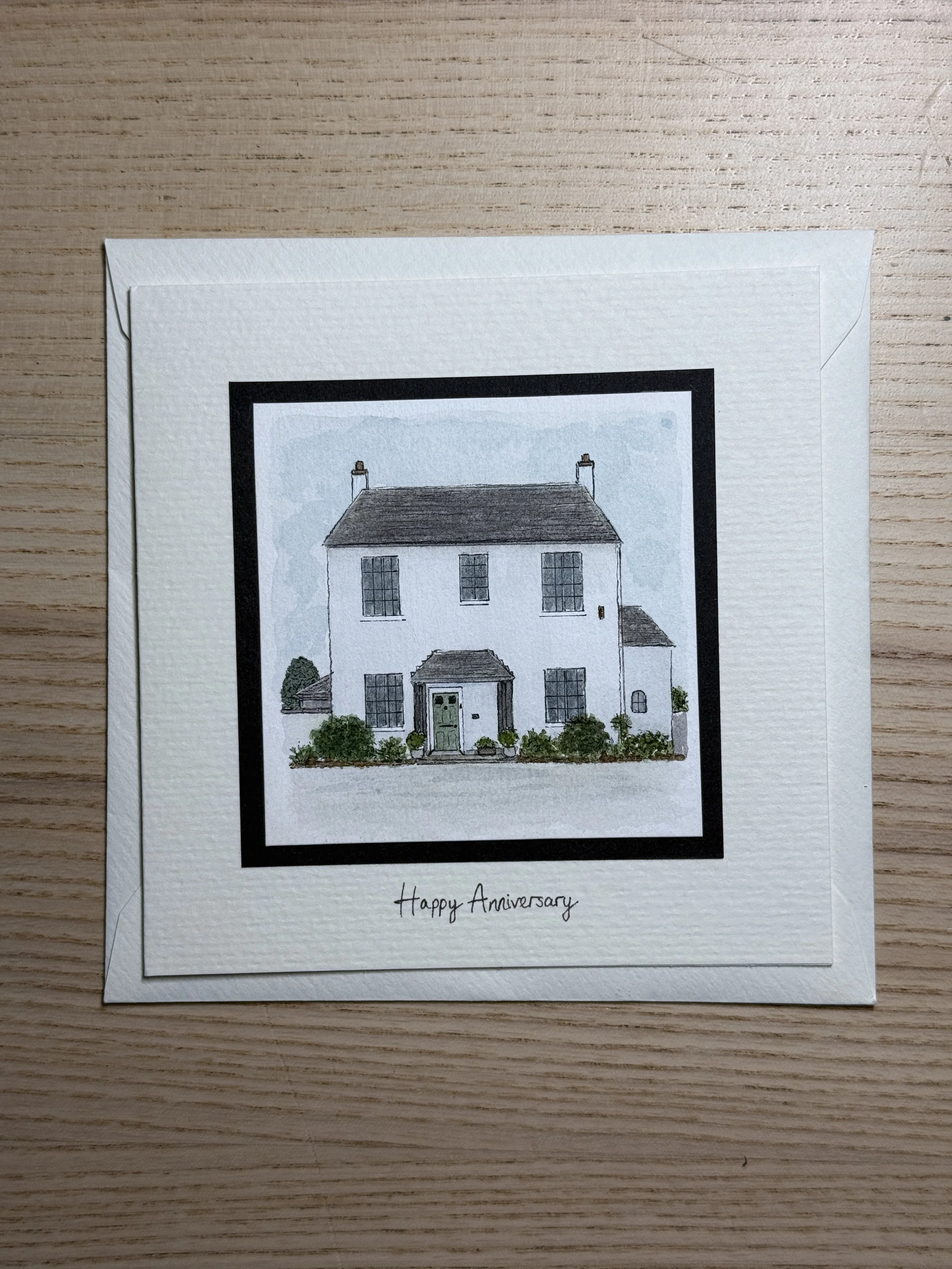 A greeting card with an illustration of a white house with a dark roof and greenery in front, and the text 'Happy Anniversary' written below the illustration.