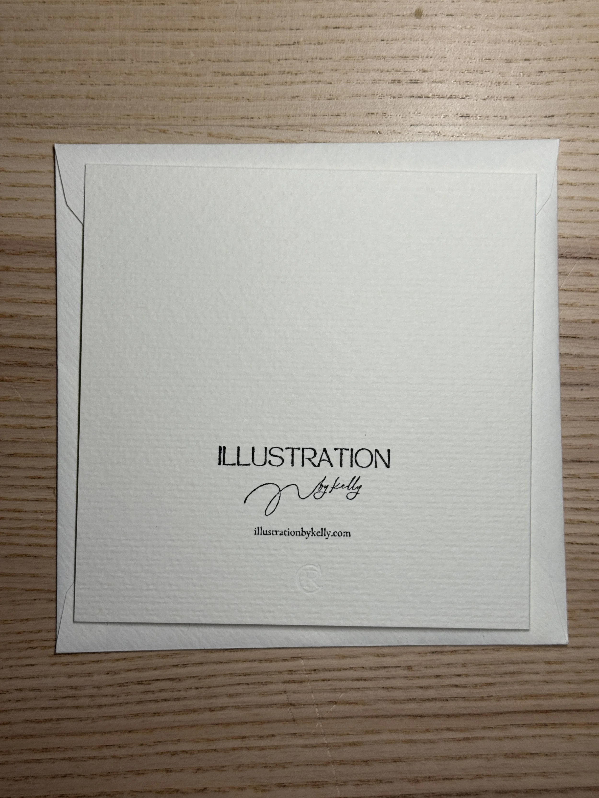 A white textured card or note with black text that reads 'ILLUSTRATION by Kelly' and a website URL 'illustrationbykelly.com', placed on a light wooden surface.