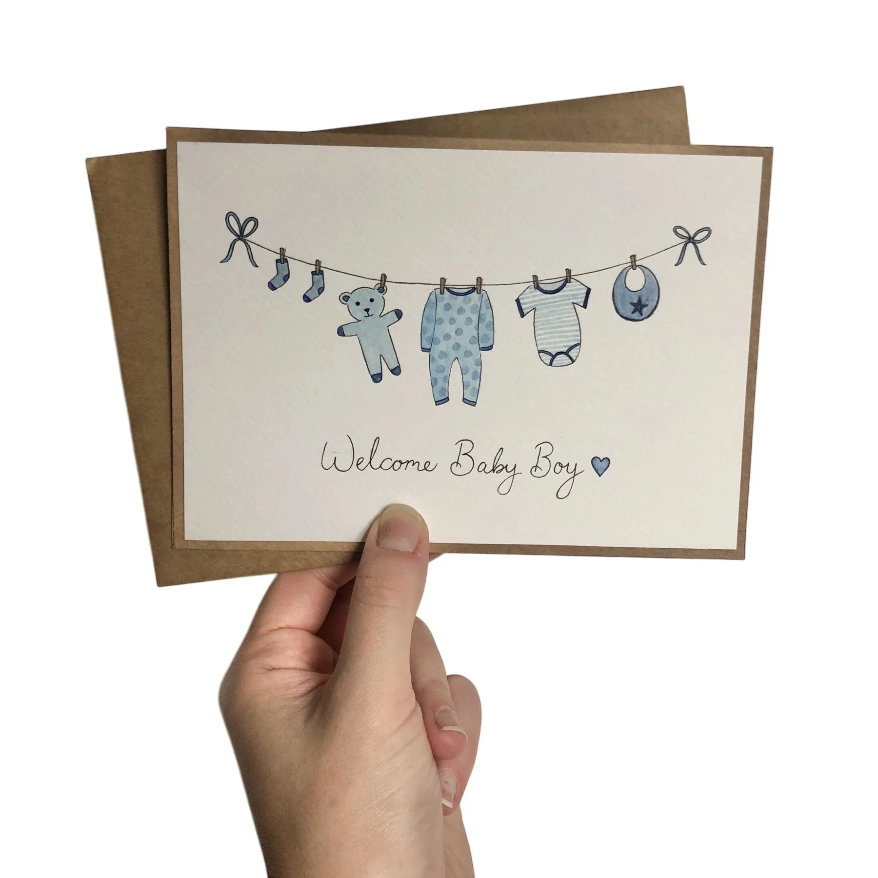 A white new baby card with a hand-drawn illustration of a clothesline with baby clothes and toys in shades of blue, and the text "Welcome Baby Boy" with a small blue heart, held by a person's hand.