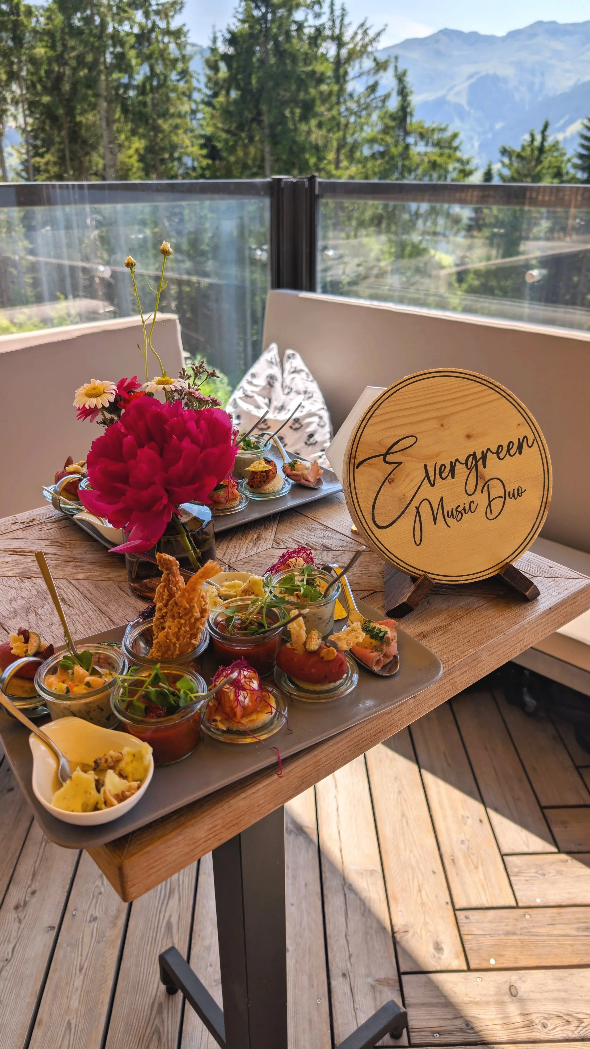 A wooden table with an assortment of appetizers in small cups, a flower arrangement with pink and yellow flowers, and a wooden sign that says "Nergreen Music Duo." The table is outdoors on a wooden deck with mountains and trees in the background.
