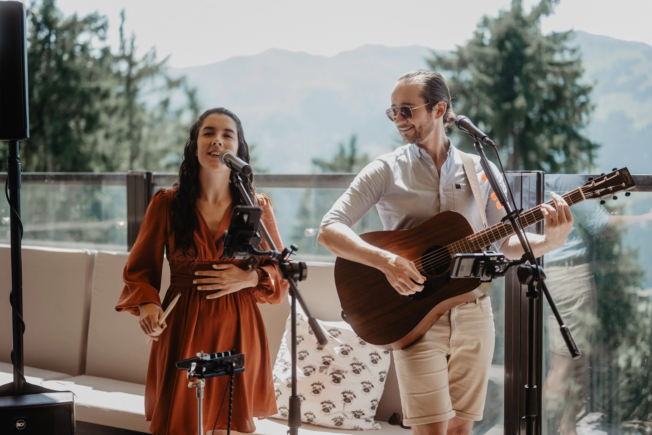 A man and woman performing music outdoors on a sunny day, with trees and mountains in the background. The woman is singing into a microphone, and the man is playing an acoustic guitar.