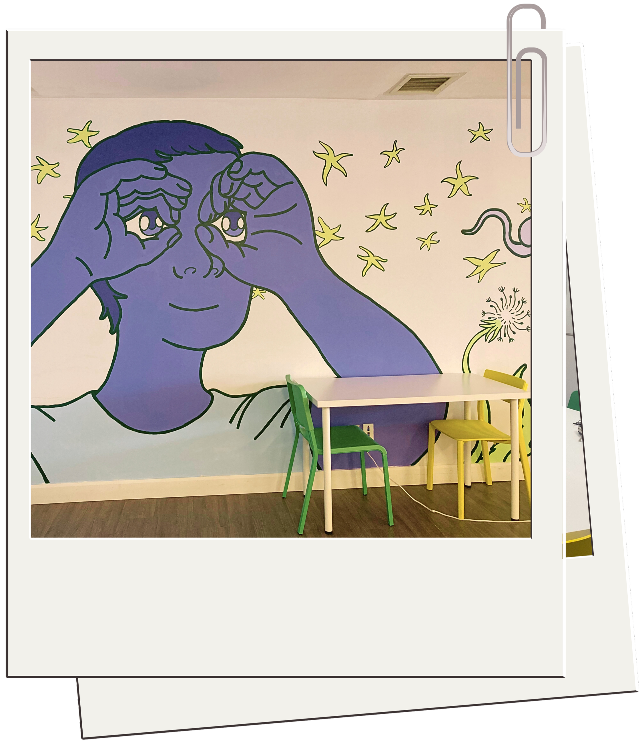 A wall mural featuring a person with blue skin making a binoculars gesture with their hands, surrounded by yellow stars and a dandelion, with a small table and green and yellow chairs in the foreground.