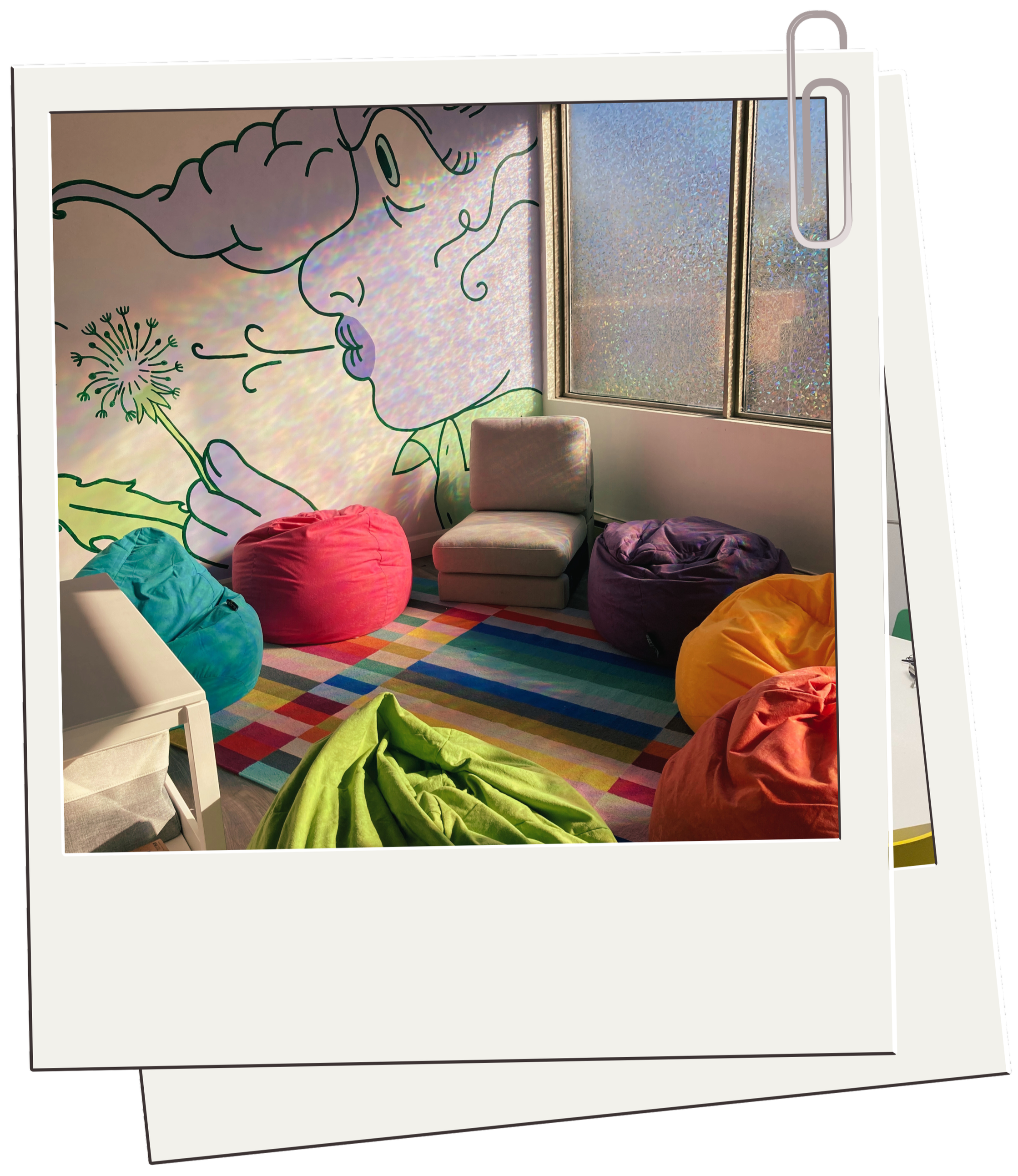 Colorful lounge area with bean bag chairs, a green blanket on the floor, a striped rug, a white armchair, and a large wall mural of a woman blowing dandelion seeds near a window with frosted glass.