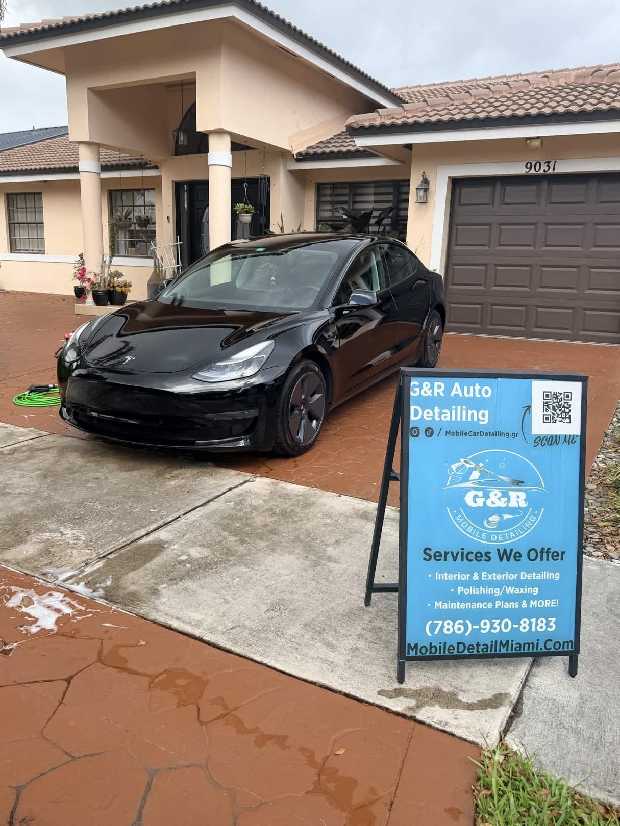 Black Tesla car parked in front of a house with a signboard for G&R Auto Detailing, offering interior and exterior cleaning, polishing, waxing, and maintenance services.