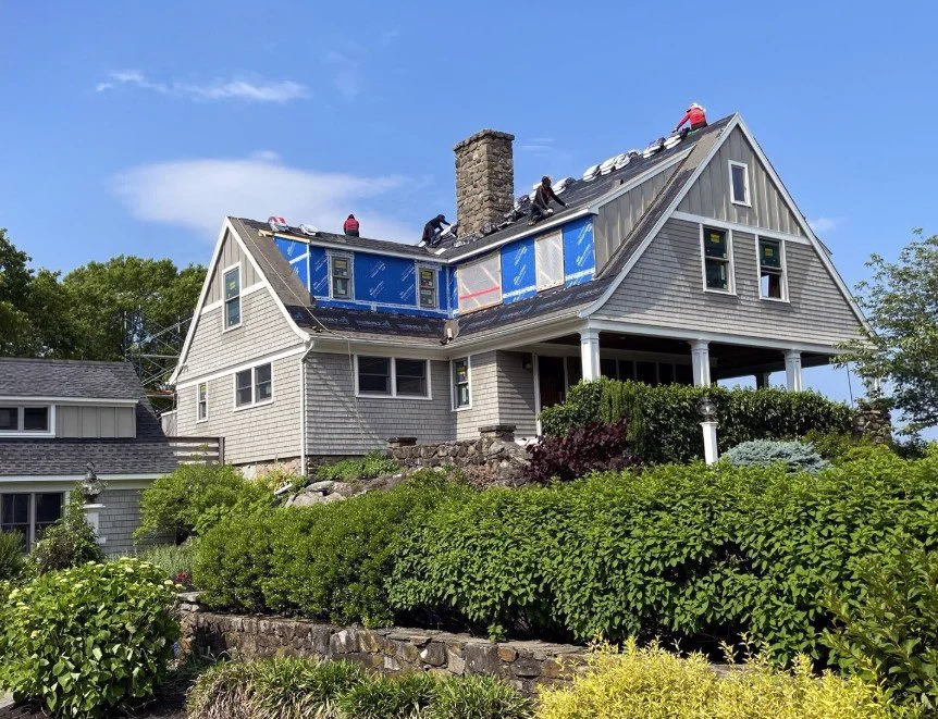 House under roof renovation with workers on the roof, blue weather-resistant barrier installed, and various roofing materials. Surrounding garden with green bushes and trees under a clear blue sky. A recent renovation project in Saunderstown RI