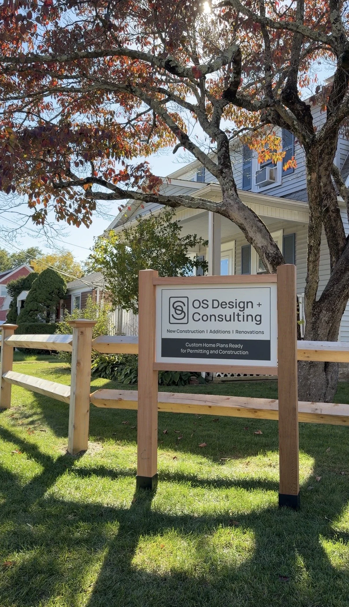 A sign for OS Design + Consulting in front of a house with a large tree, featuring fall foliage and a wooden fence. The sign indicates services like new construction, additions, renovations, and custom home plans.
