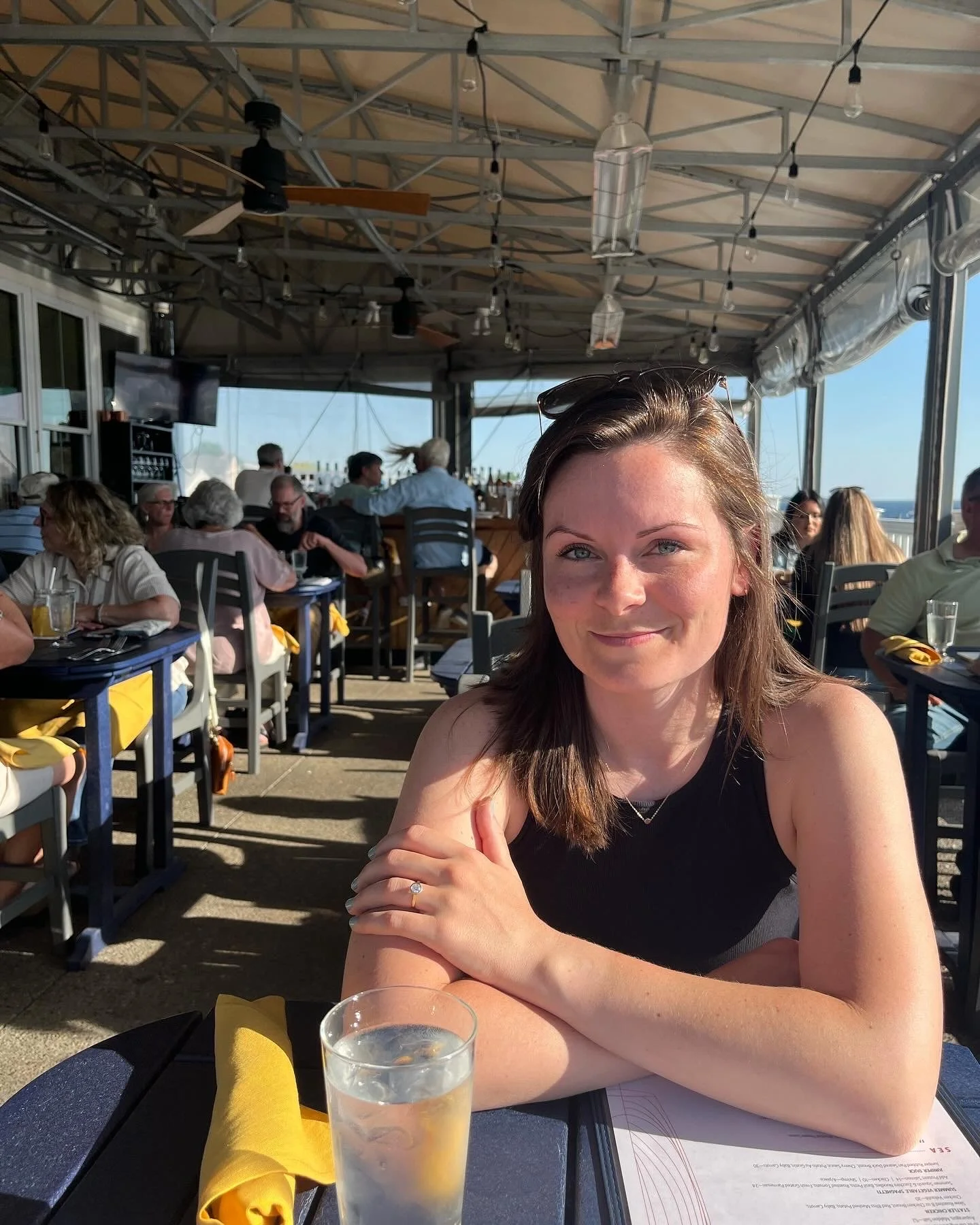A woman sitting at a restaurant table with a glass of water, smiling at the camera. The restaurant has an open-air setup with a view of the ocean in the background and other diners seated around.