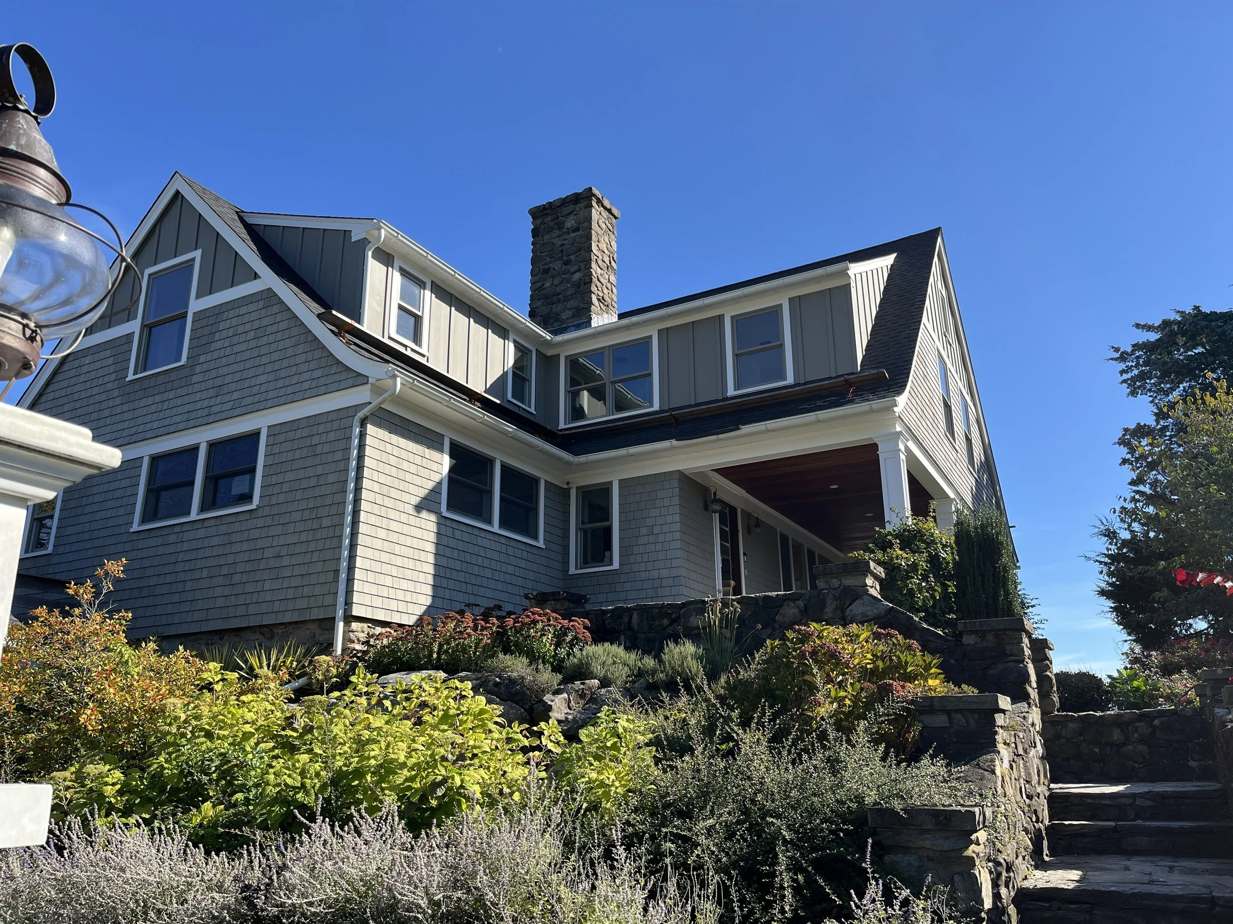 A multi-story house with gray and beige siding, a stone chimney, and a garden with various plants and flowers in the foreground under a clear blue sky in Saunderstown Rhode Island. The home was a recent renovation project