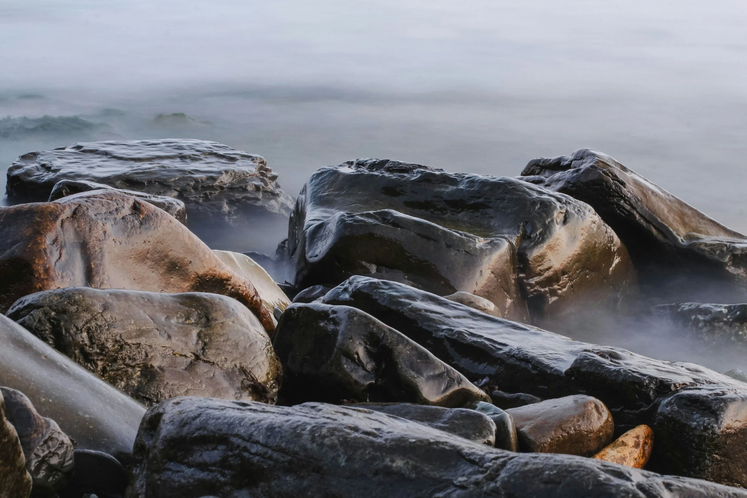 Smooth, wet rocks along a shoreline with water washing over them in the background.