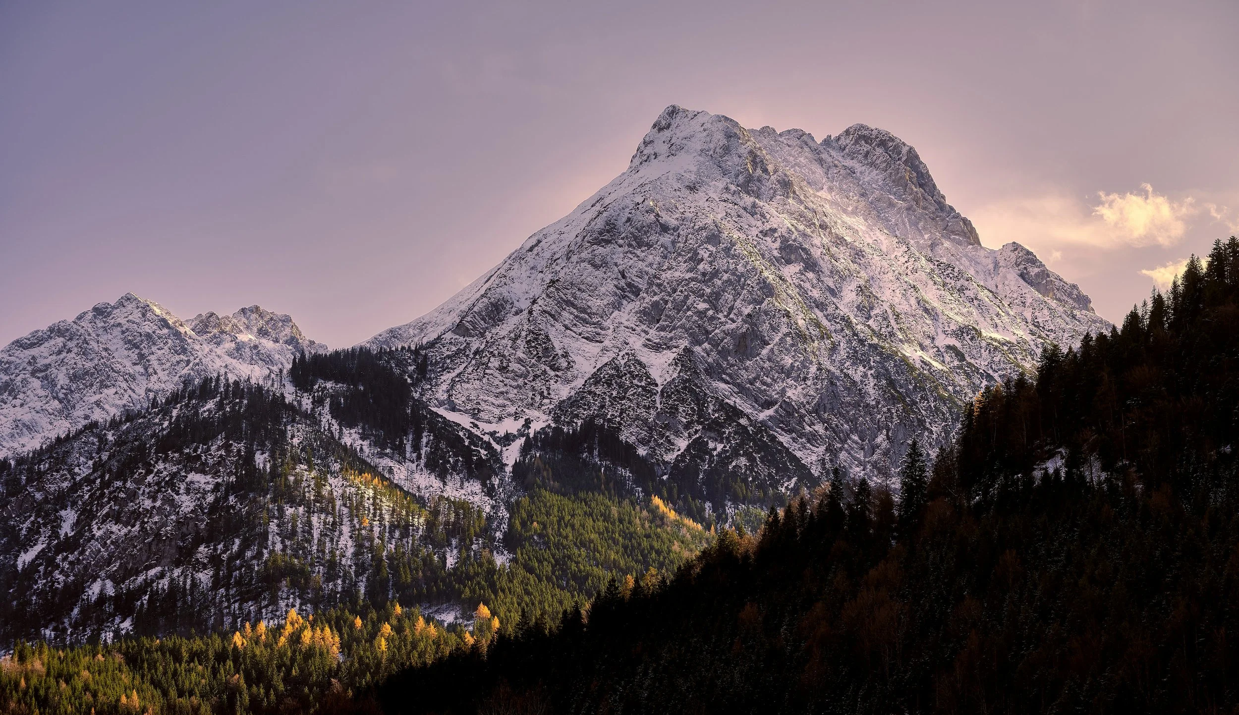 Snow-covered mountain with a forested base and a partially cloudy sky during sunset.