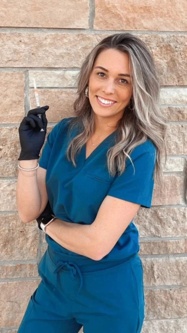 A woman in blue scrubs holding a syringe in front of a brick wall, smiling.