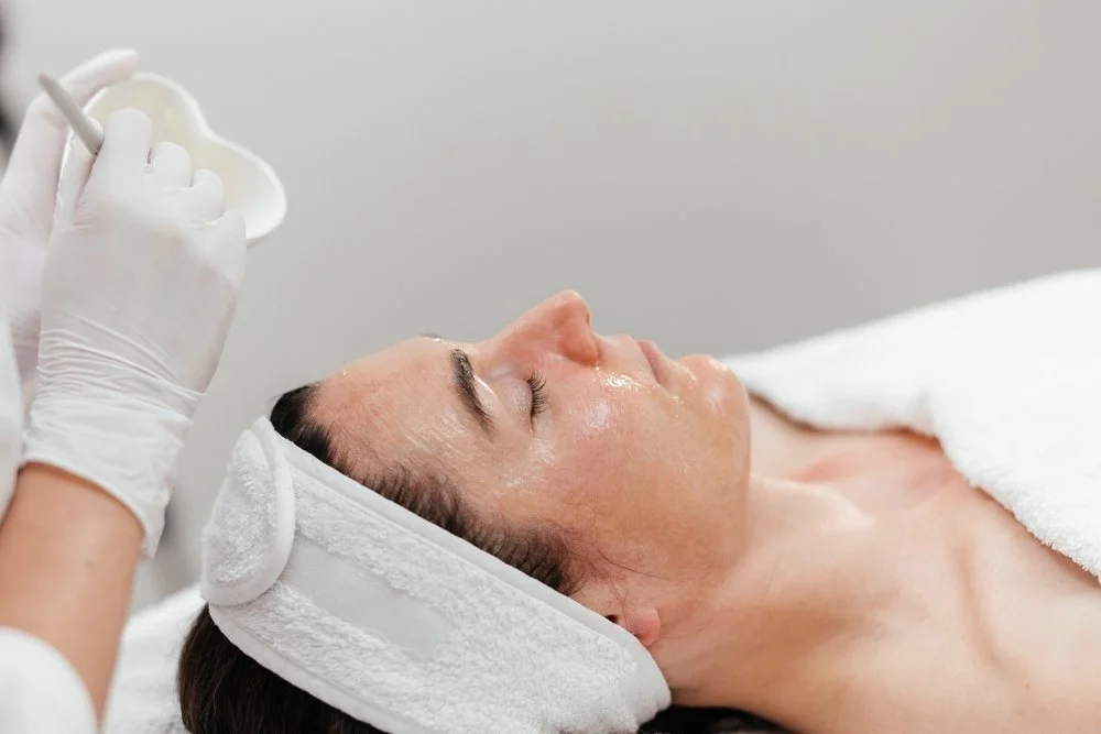 A woman lying on a spa bed with a towel wrapped around her head, receiving a facial treatment from a skincare professional wearing gloves.