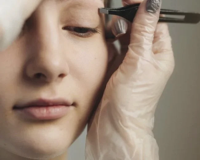 Close-up of a person getting an eyebrow waxing or plucking, with a beautician wearing gloves holding tweezers near their eyebrow.
