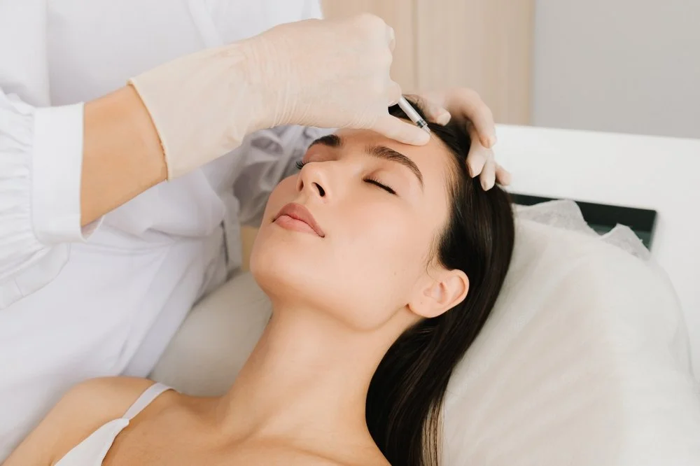 A woman receiving a cosmetic injection in her forehead at a clinic, lying down with her eyes closed.