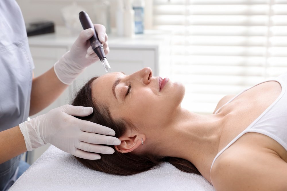 A woman lying on a treatment bed receiving a cosmetic procedure with a professional using a pen-like device.