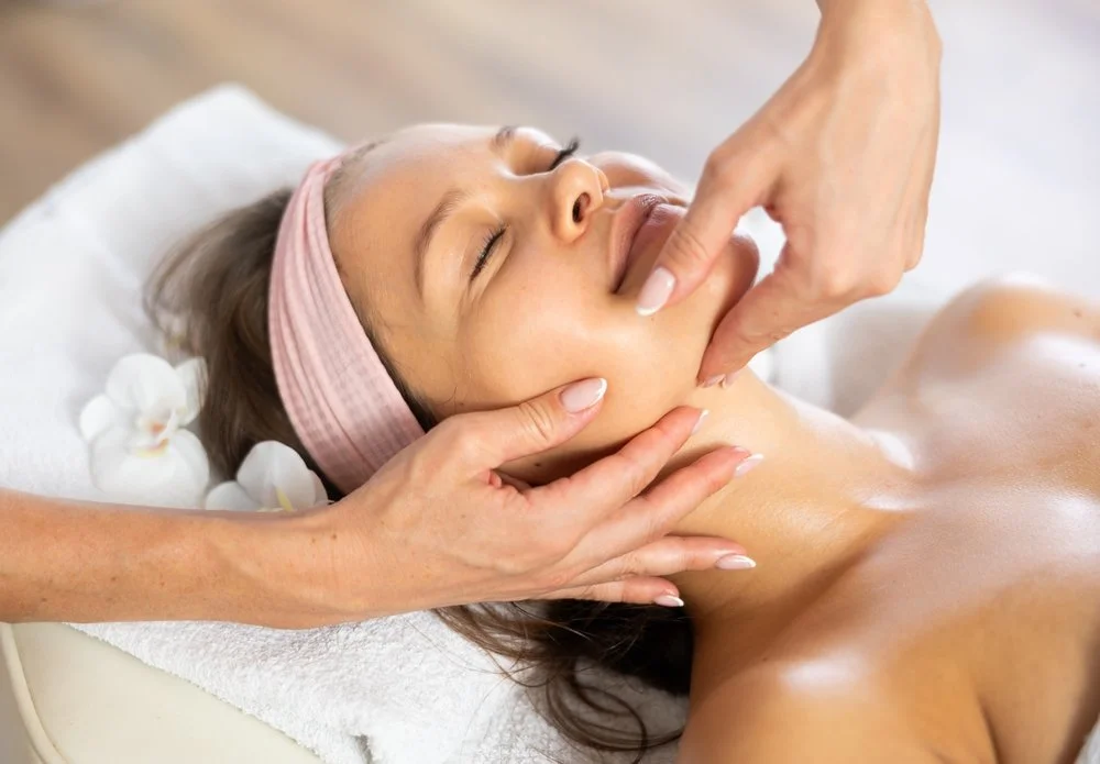 A woman receiving a facial massage at a spa, lying on a treatment bed with flowers in her hair and a pink headband, eyes closed.