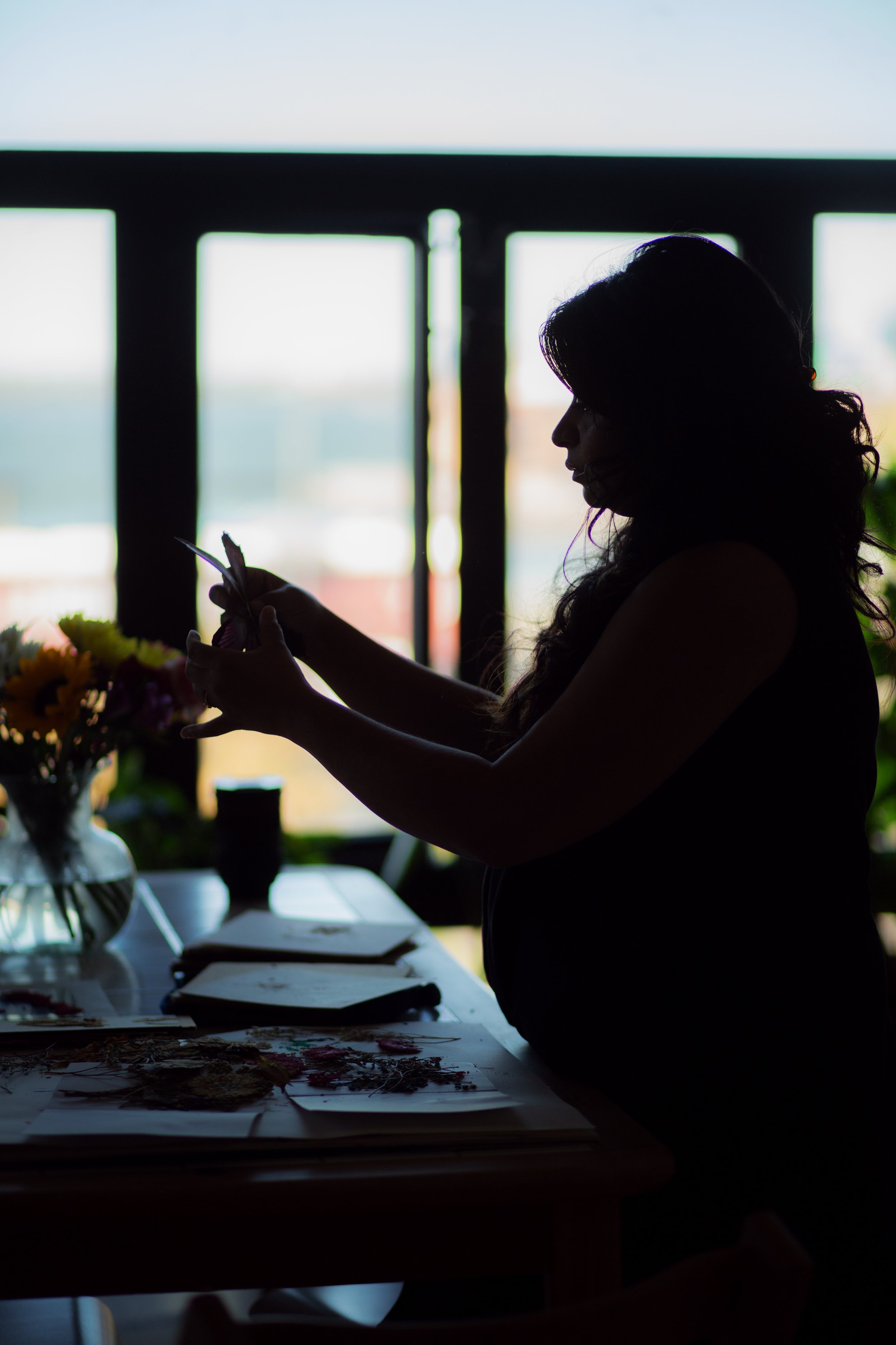 Silhouette of a woman sitting at a table, working on an art project with flowers and papers, near a window with bright light outside.