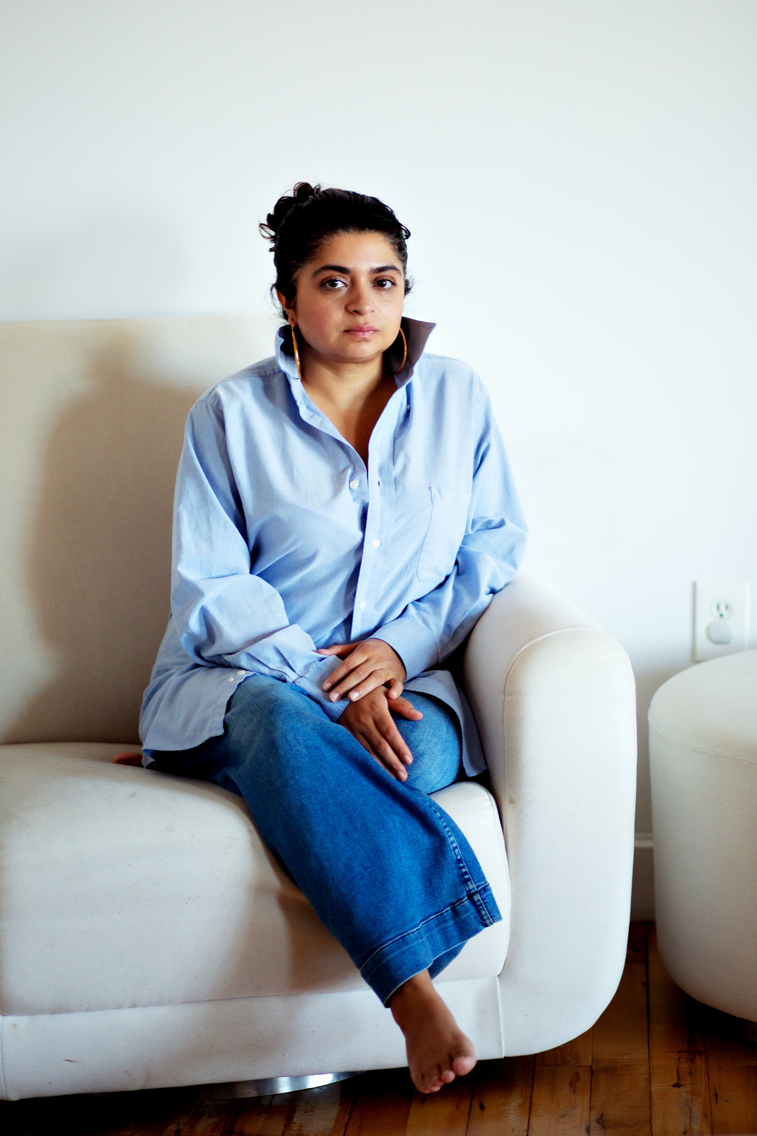 A woman sitting on a white sofa, wearing a light blue shirt and jeans, with one foot on the sofa and the other hanging down, in a minimalist room with a wooden floor and a white wall.