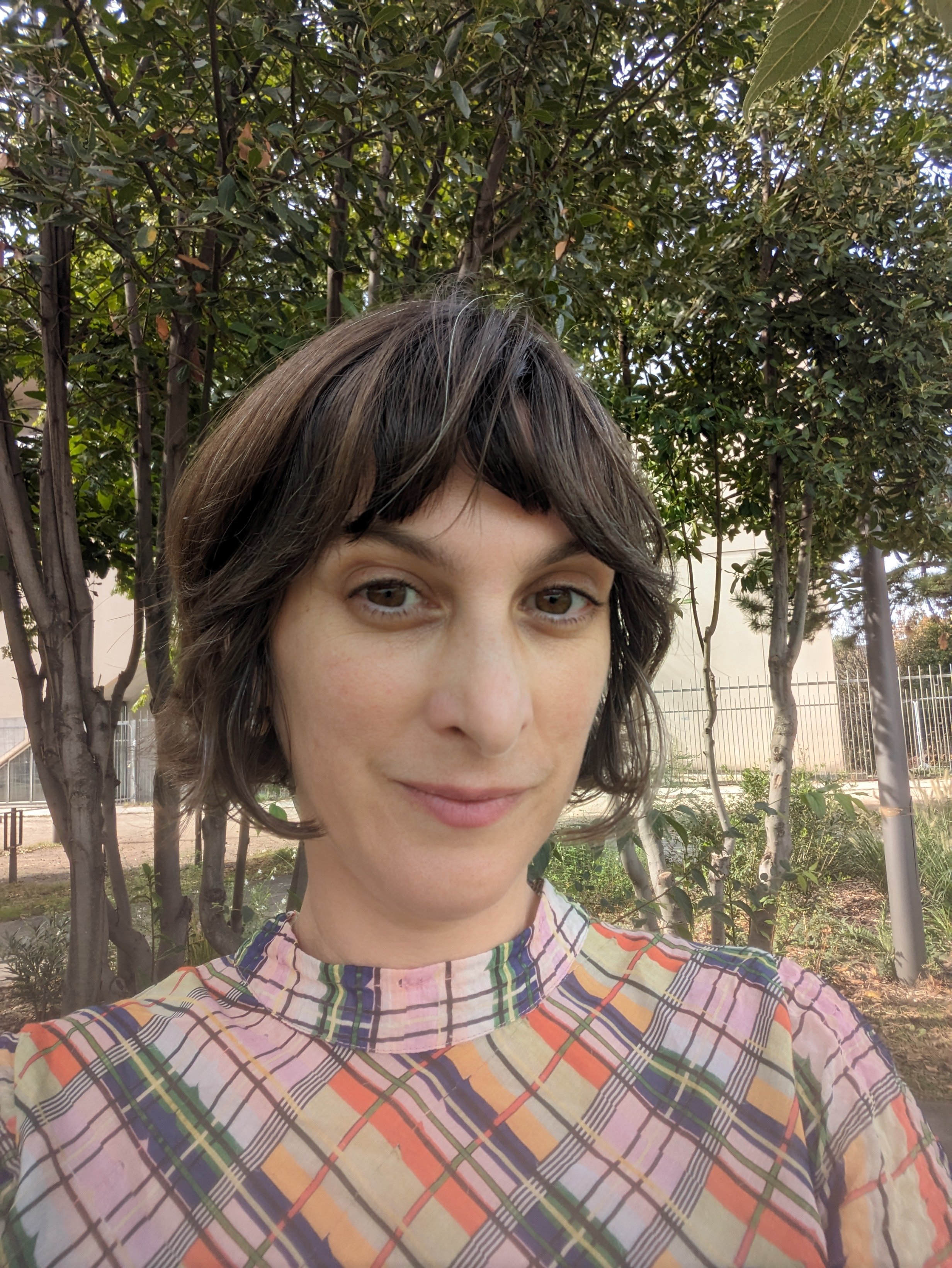 Close-up of a young woman with short brown hair, wearing a colorful plaid shirt, standing outdoors in front of trees and a fence.