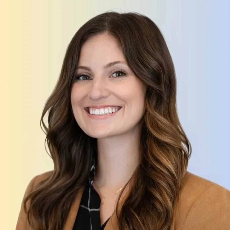 Portrait of a smiling woman with long, wavy brown hair and light makeup wearing a tan blazer and black top, with a colorful gradient background.
