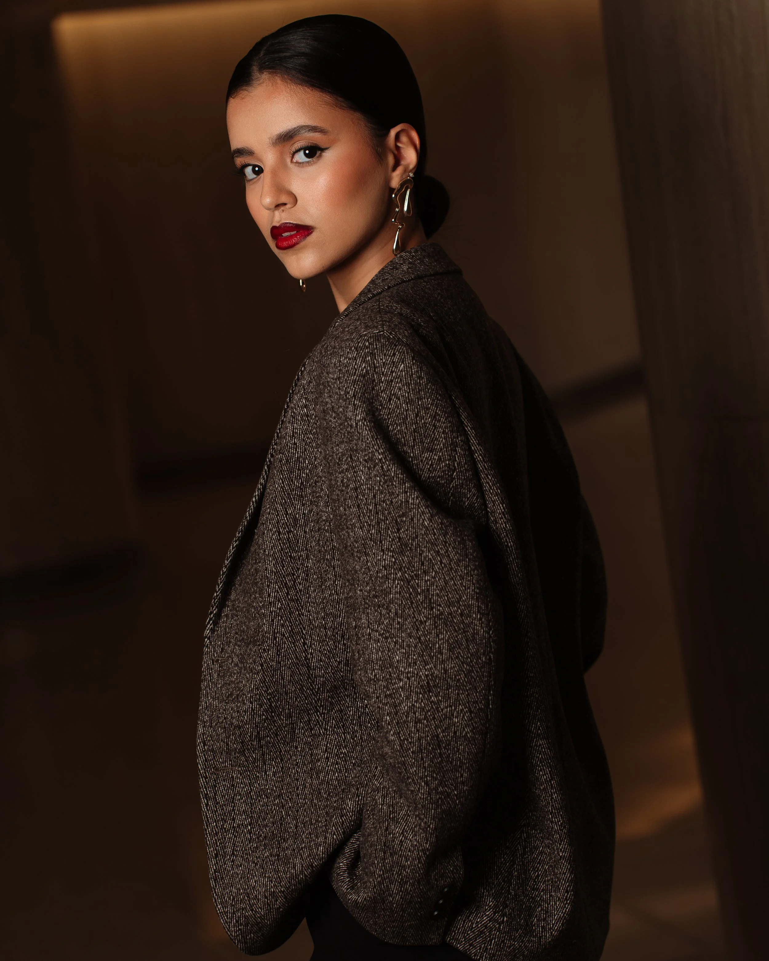 A young woman with dark hair tied back, wearing bold red lipstick, gold earrings, and a brown patterned blazer, looking over her shoulder in a dimly lit indoor setting.