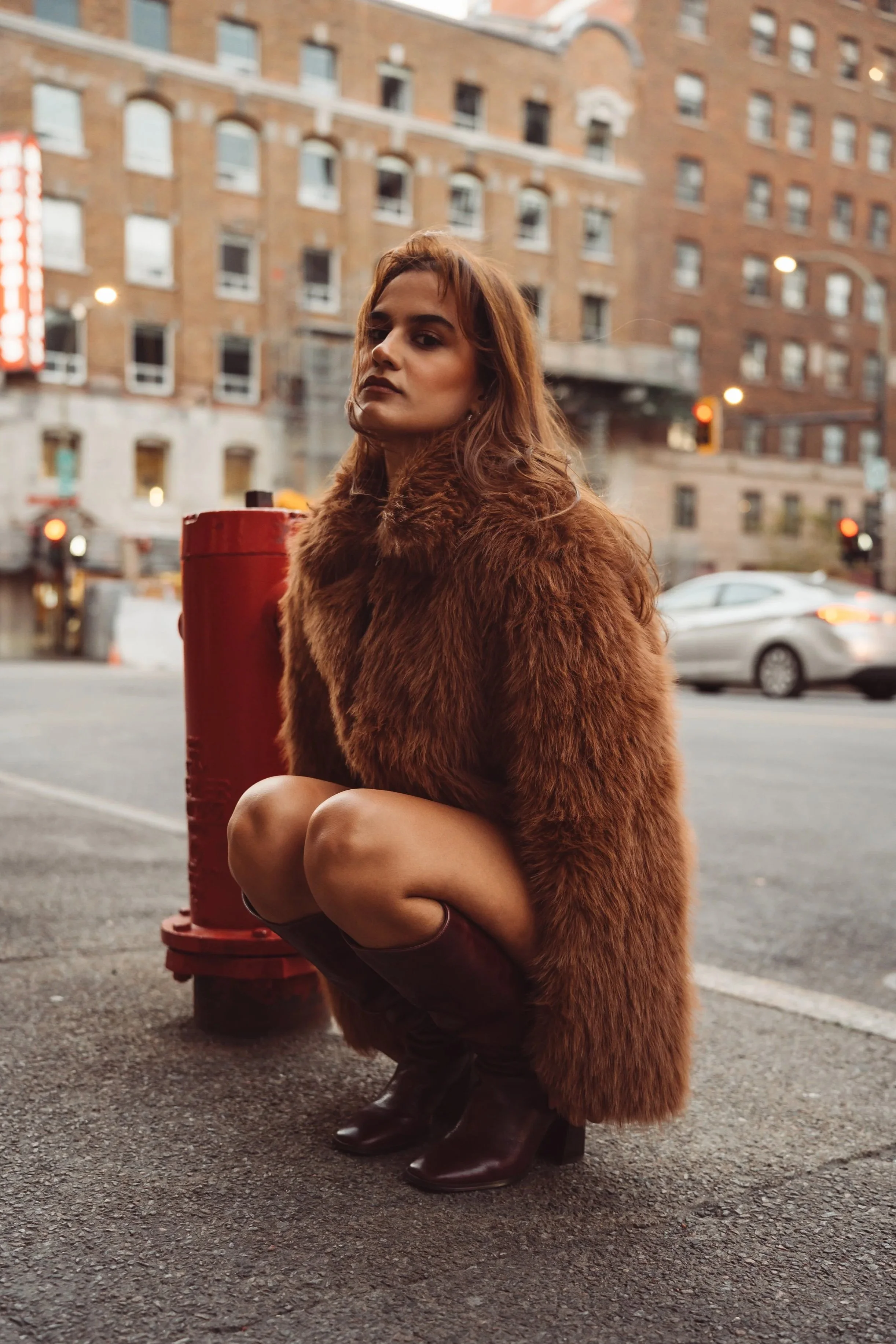 A woman with long brown hair and a serious expression squats on the city street, wearing a brown fur coat and knee-high boots, with a cityscape background of old brick buildings and moving cars.