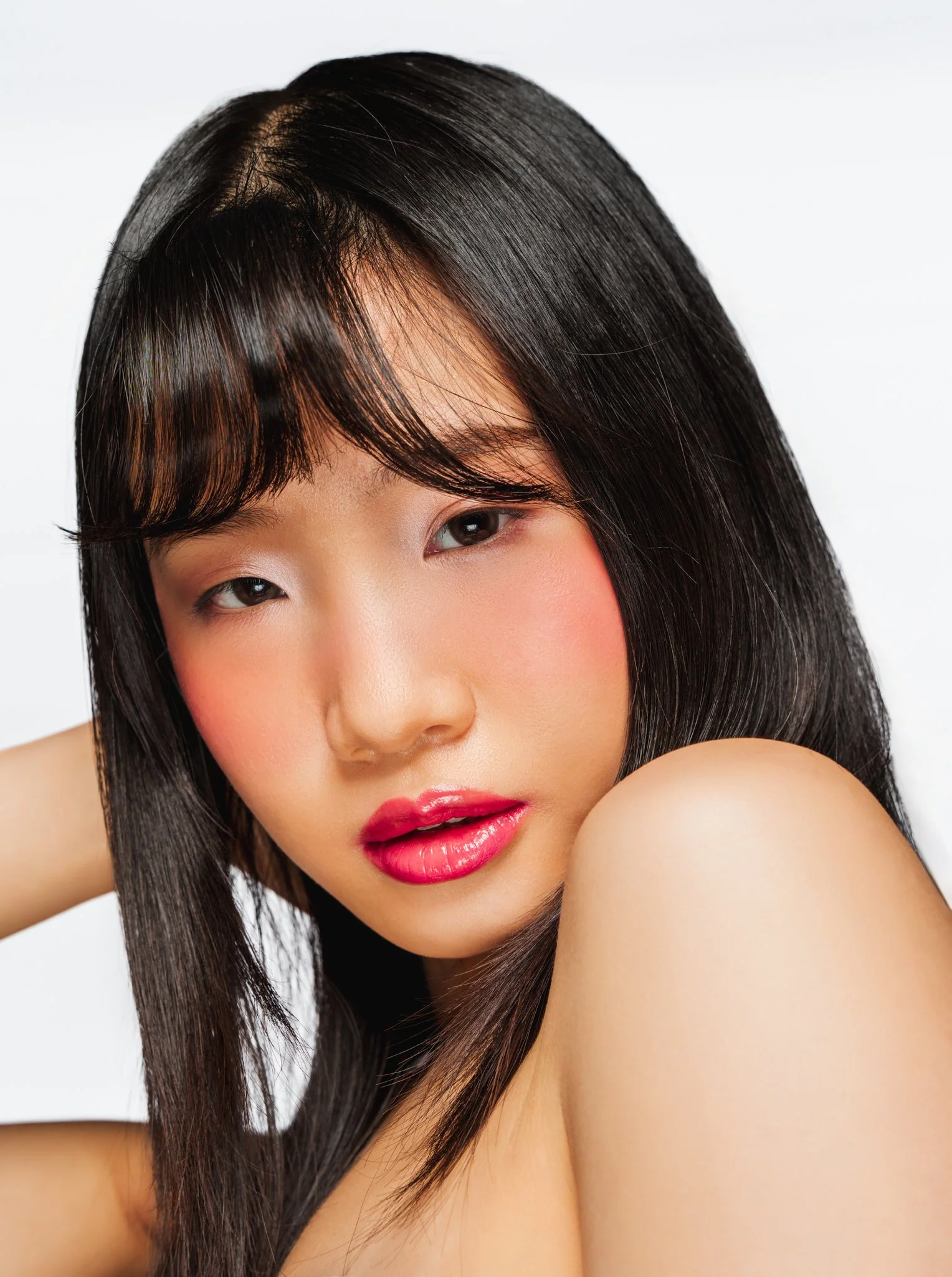Close-up of a woman with long black hair, glossy pink lips, and light makeup, posing with her shoulder near her face against a white background.