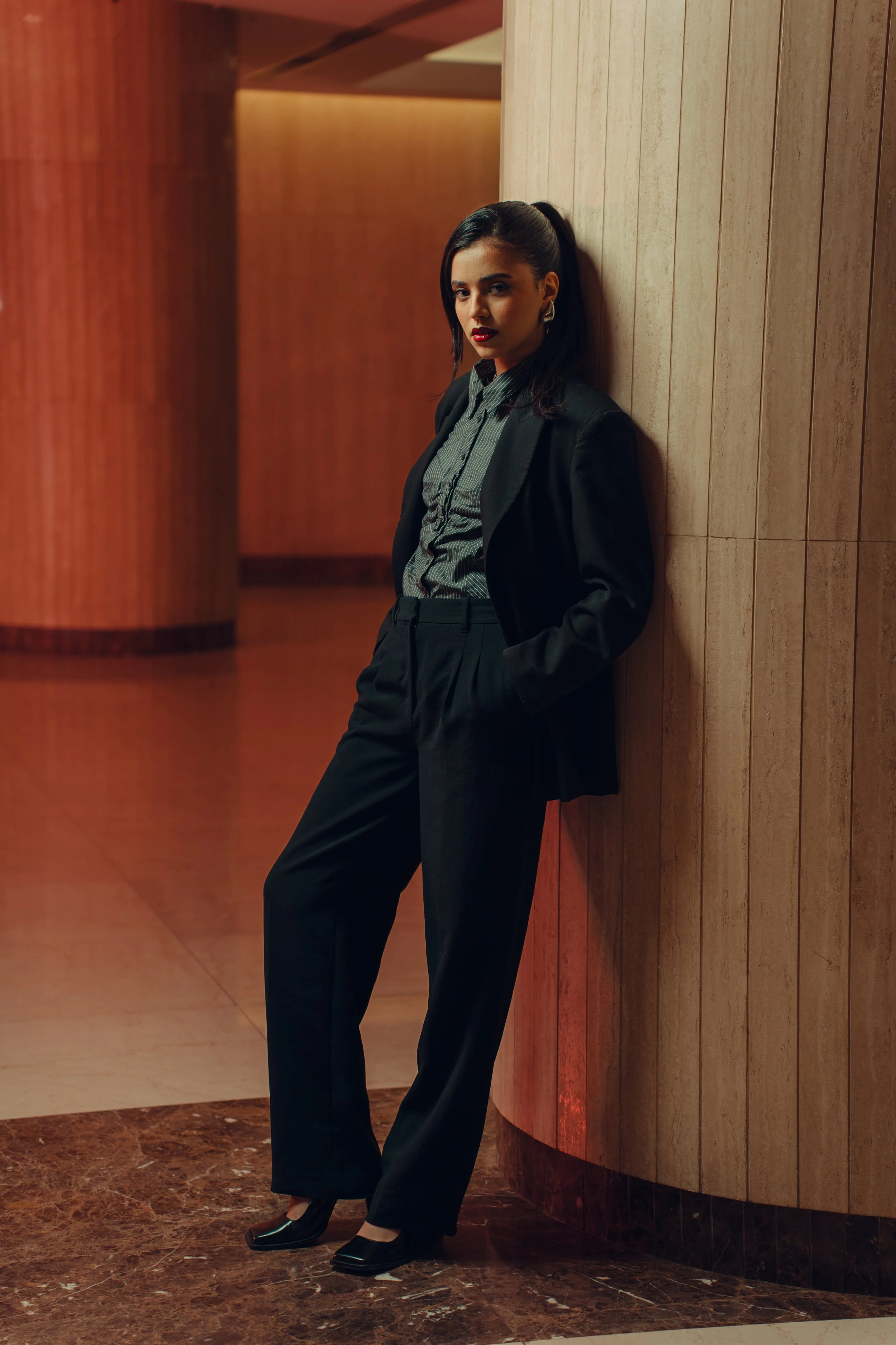 A woman in a black suit and heels leaning against a wooden wall, in a dimly lit, modern indoor setting.