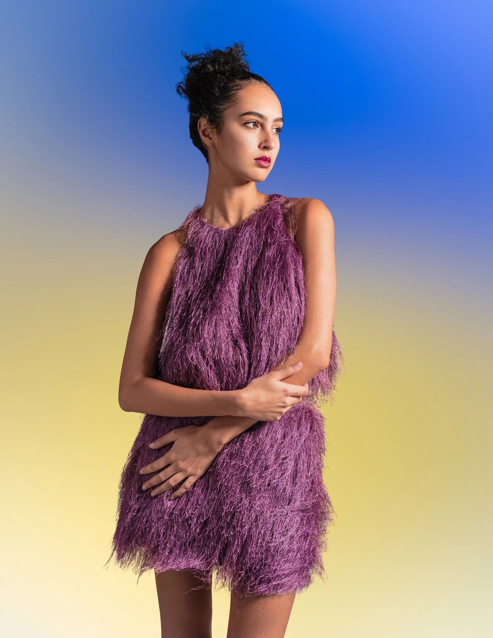 A young woman with curly black hair in a high bun, wearing a sleeveless purple feathered dress, stands with her arms crossed against a blue and yellow gradient background.