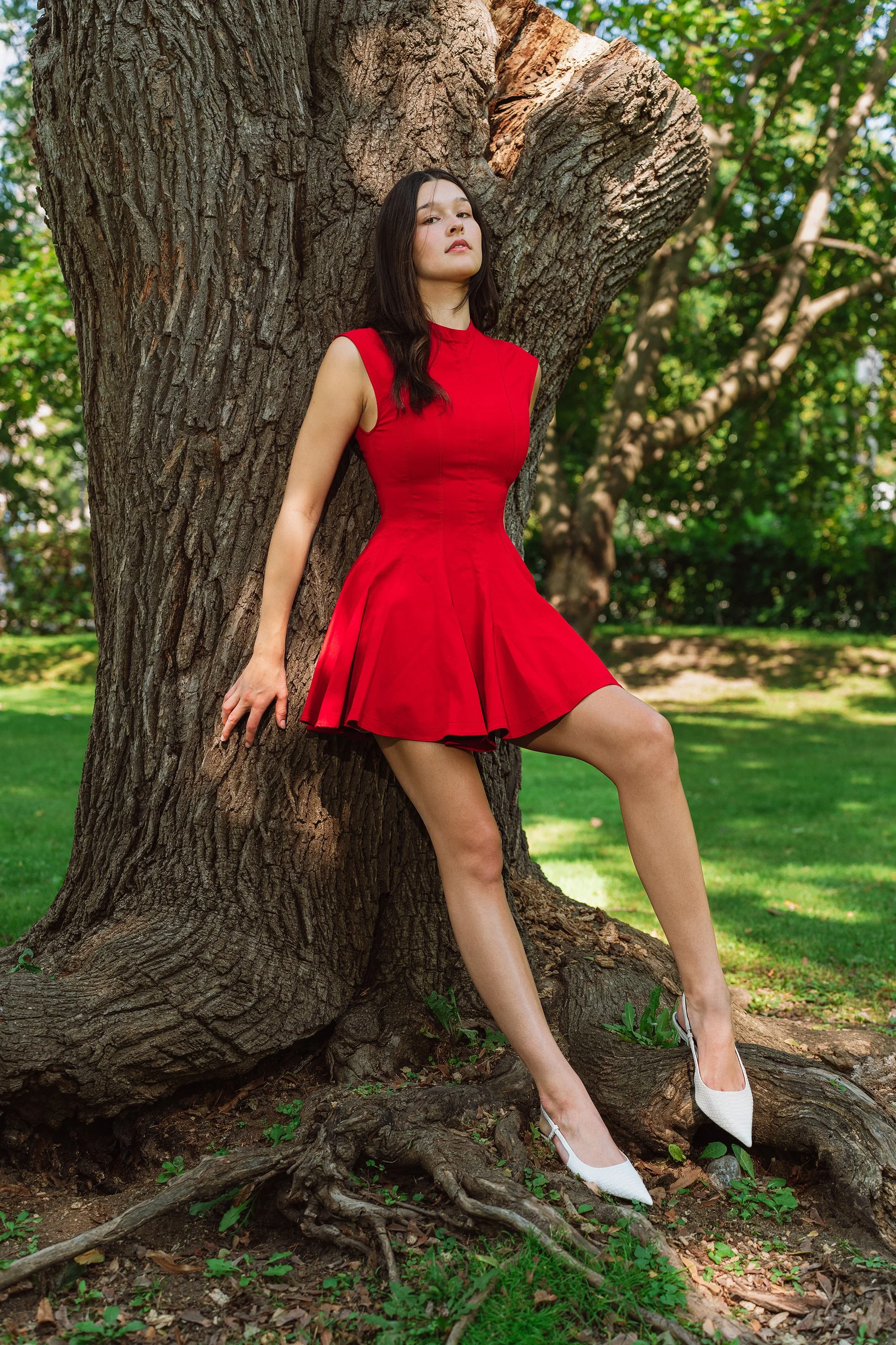 A woman in a red sleeveless dress and white high heels leaning against a large tree in a park with green grass and trees in the background.