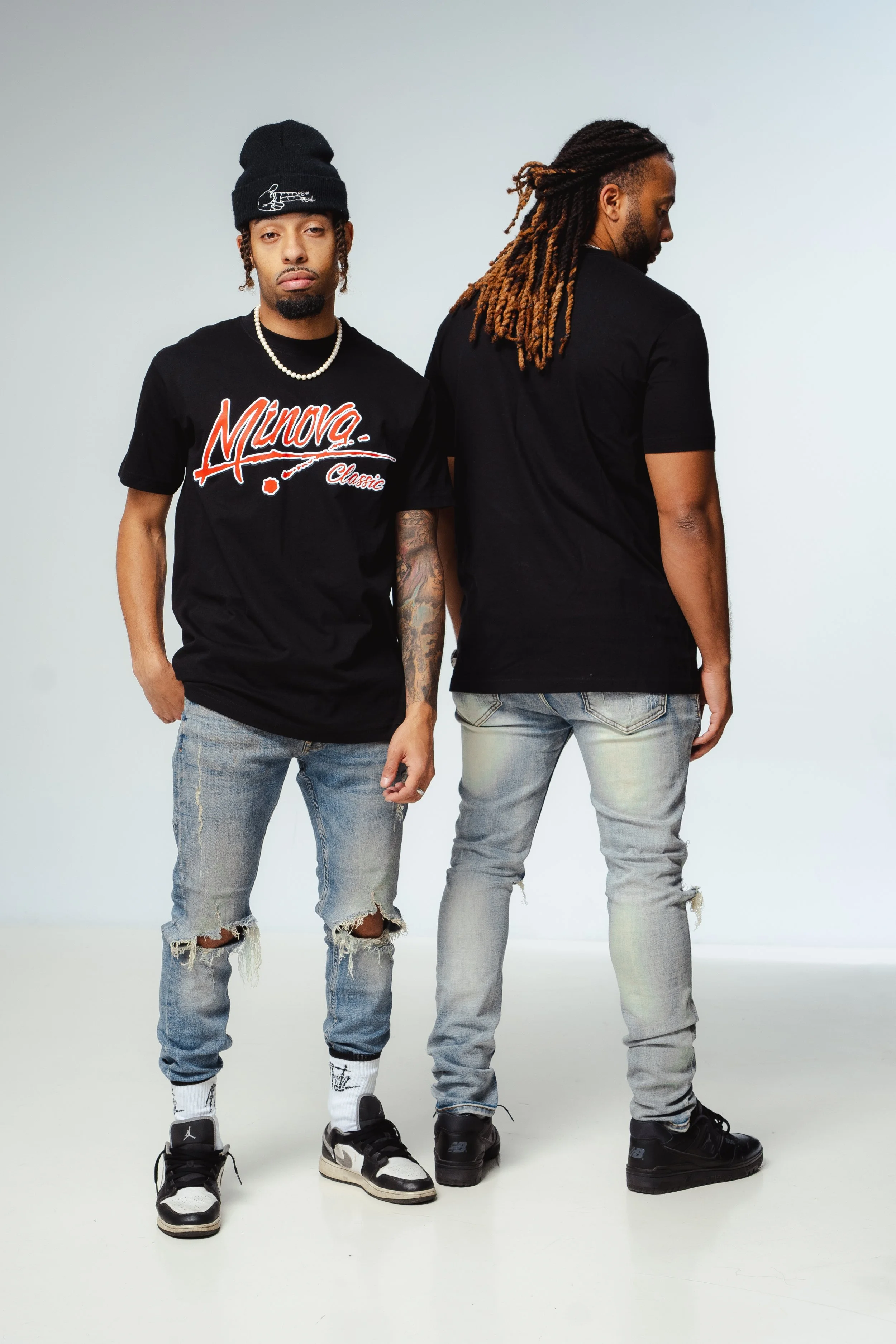Two men standing against a light background, one facing forward with hands in his pockets and the other turned to the side, wearing casual streetwear including T-shirts, ripped jeans, and sneakers.