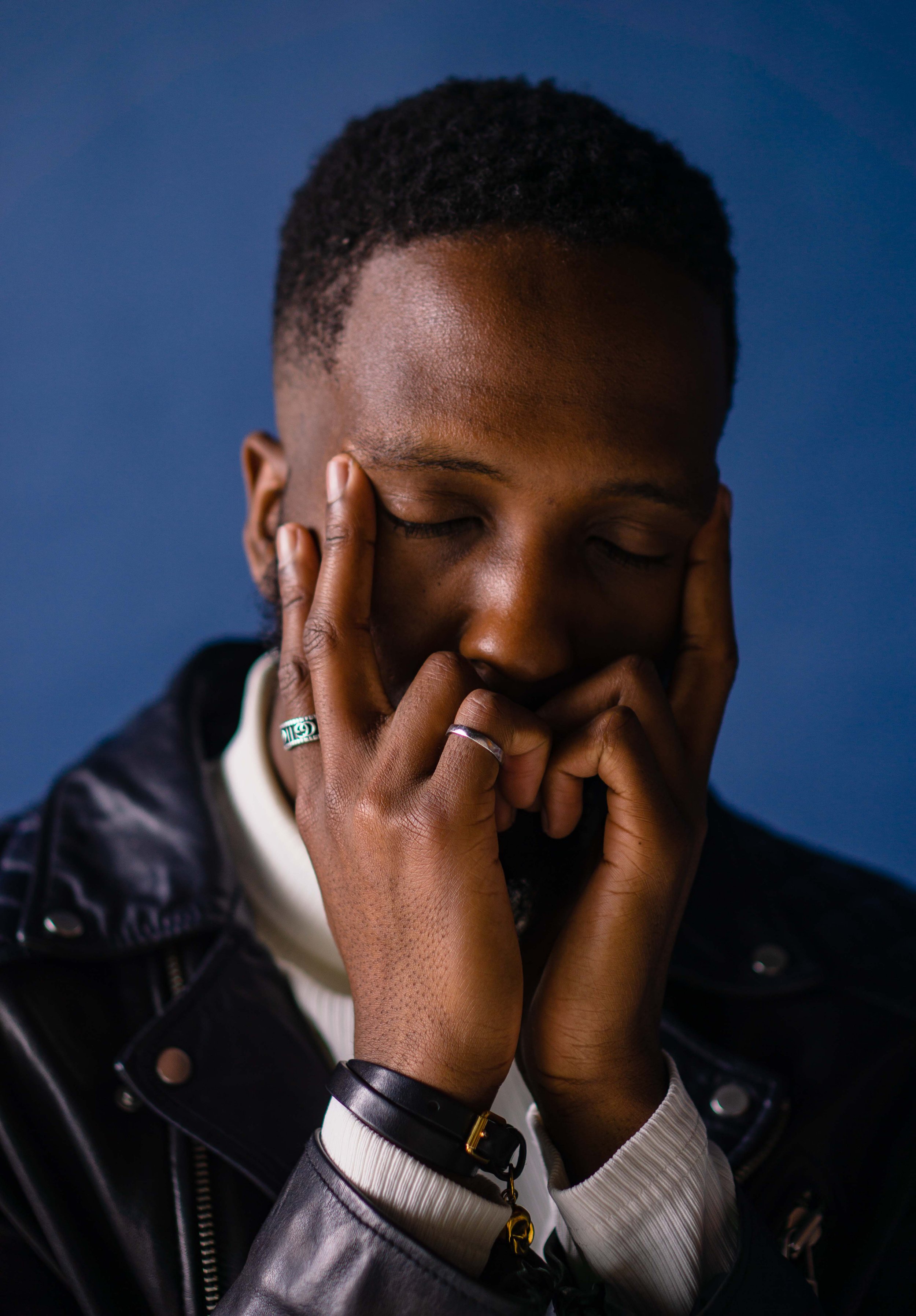 A young man with short curly hair and dark skin, eyes closed, resting his face in his hands, wearing a black leather jacket, with rings on his fingers and a black bracelet on his wrist, against a dark blue background.