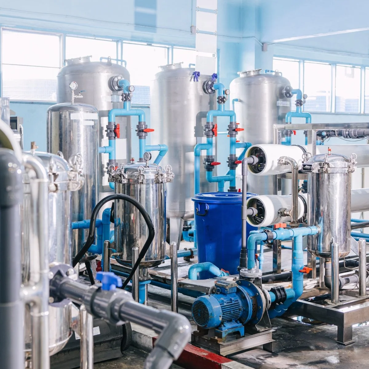 Industrial water treatment equipment with stainless steel tanks, blue pipelines, gauges, and a blue filter housing in a factory setting.