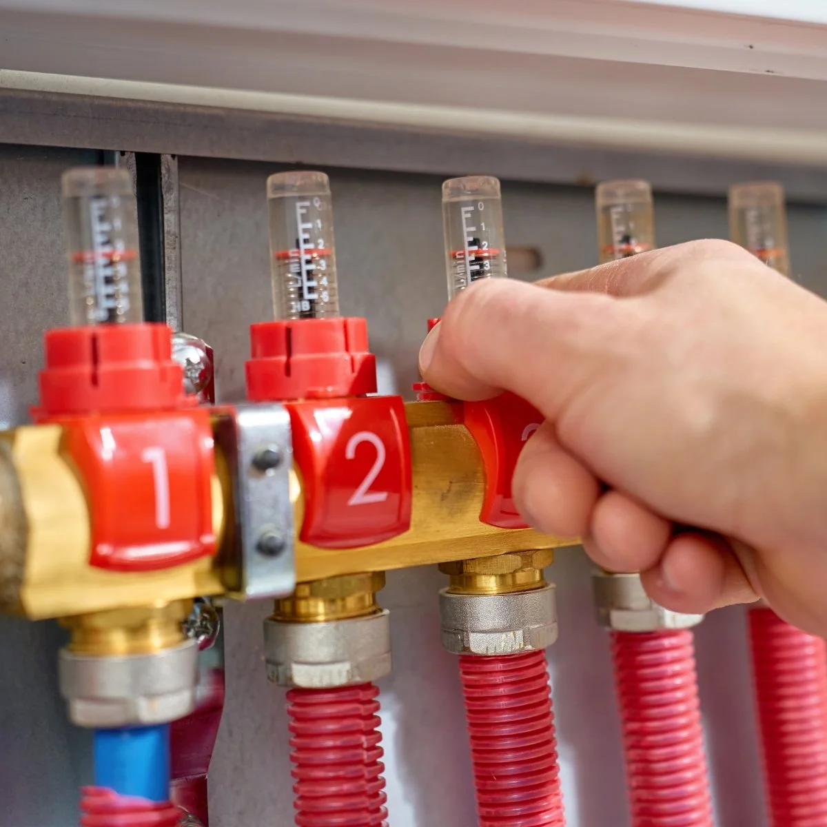 A person's hand adjusting a red valve on a plumbing manifold with numbered indicators, connected to colored pipes.
