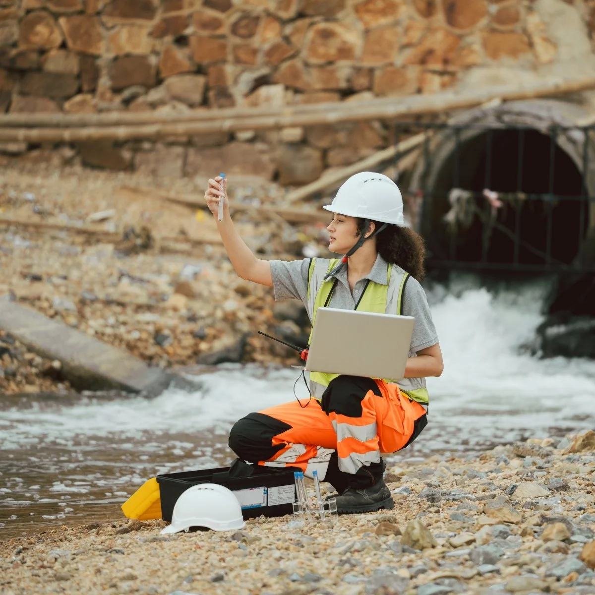 A female engineer wearing a hard hat, safety vest, and orange work pants kneels on a rocky riverbank holding a test tube and a laptop, with a tunnel and flowing water in the background.