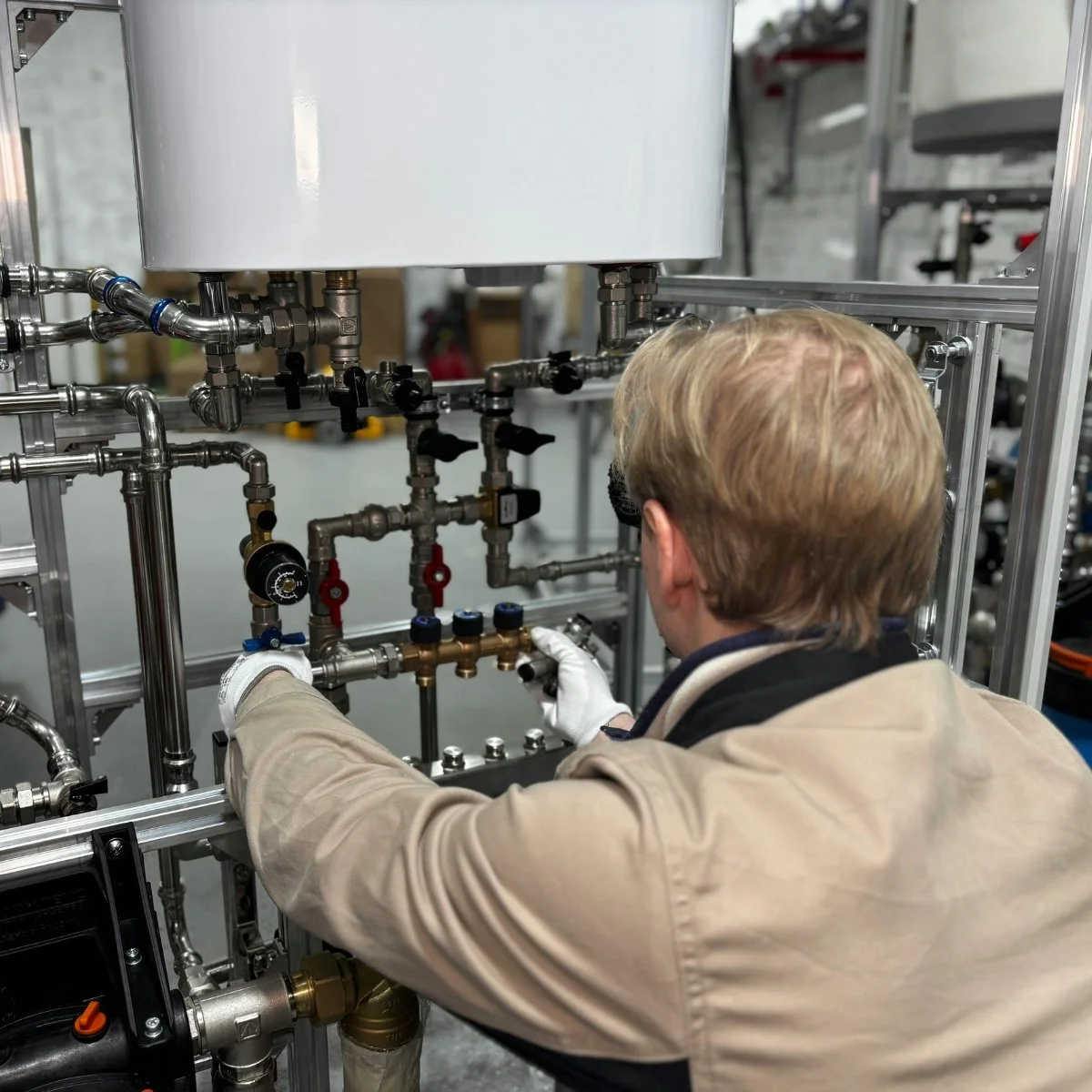 A person wearing white gloves working on a complex network of metal pipes and valves in an industrial or laboratory setting.