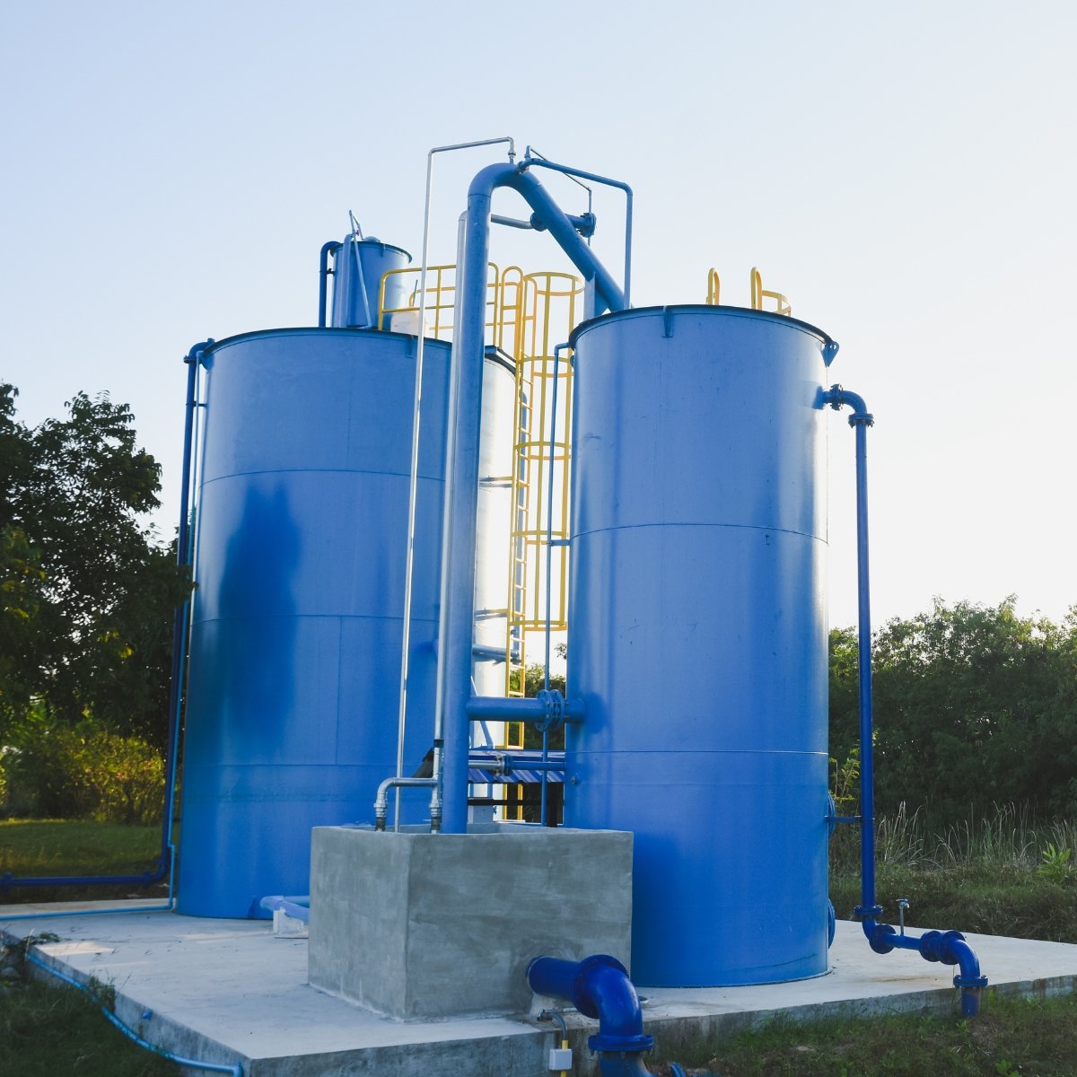 Two large blue industrial storage tanks connected by pipes, situated outdoors on a concrete platform, with trees in the background.