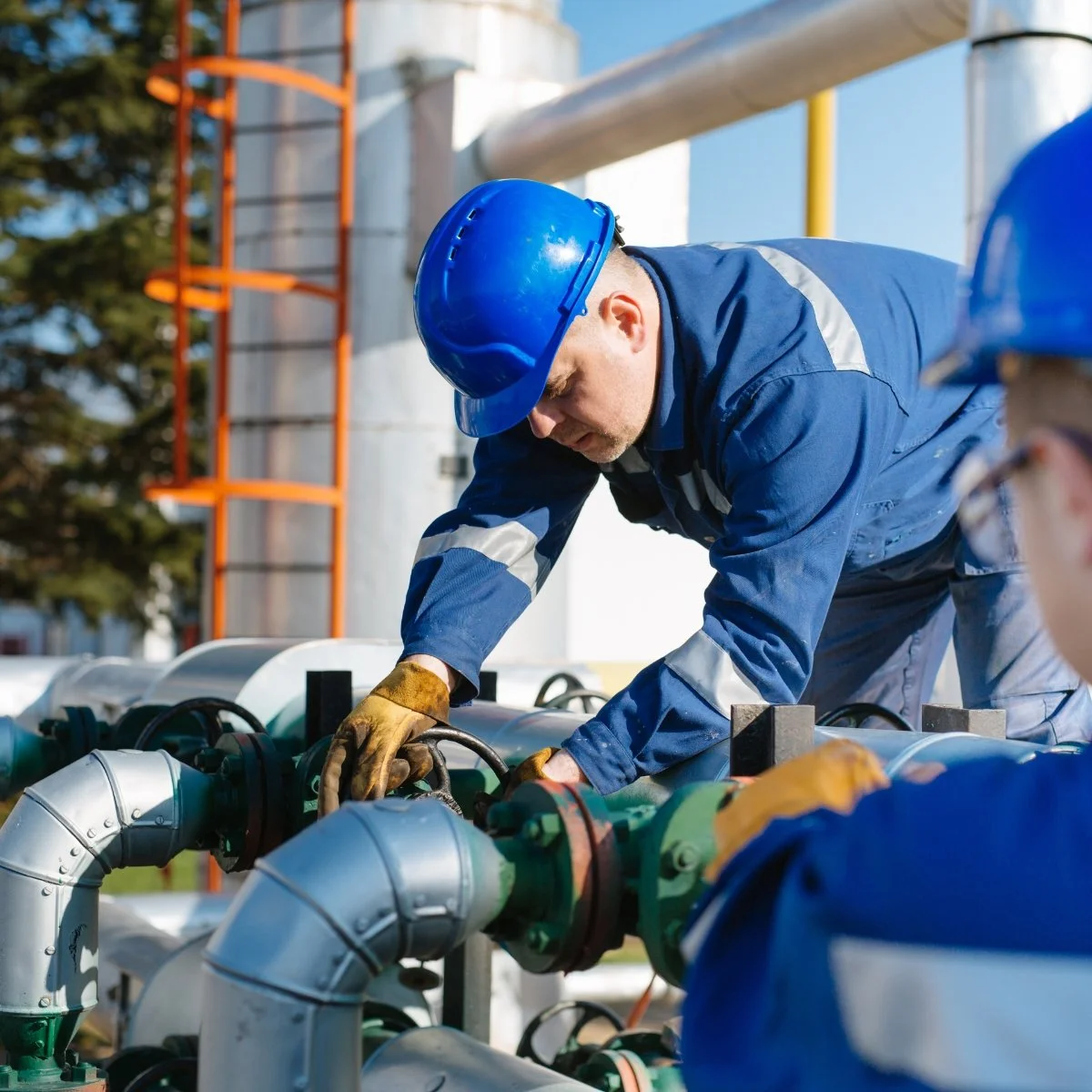 Worker in blue safety uniform and helmet inspecting pipes at an industrial site.