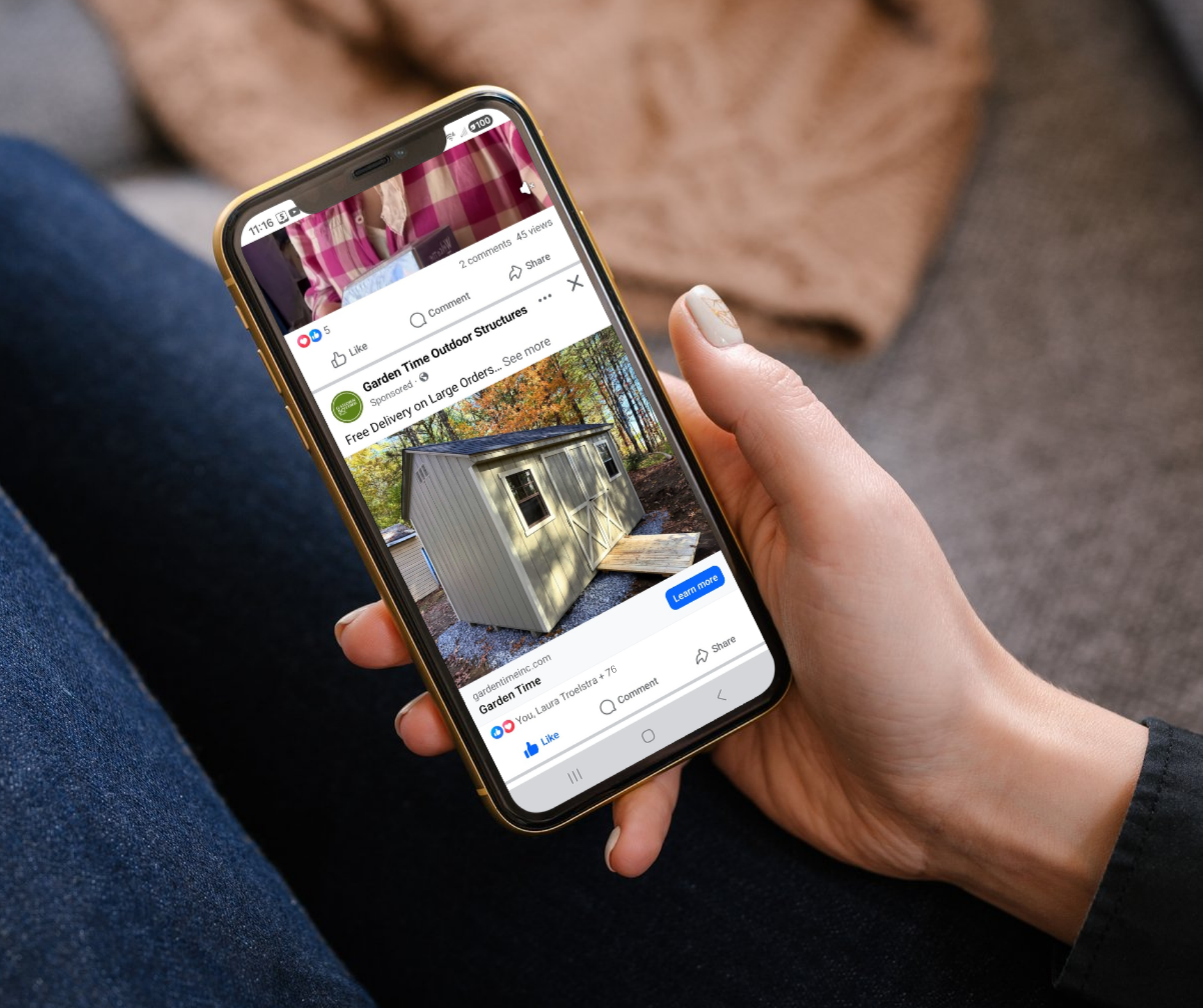Person holding a smartphone displaying a Facebook post about a small outdoor shed with a peaked roof, window, and side door, situated on a gravel surface in a wooded area.