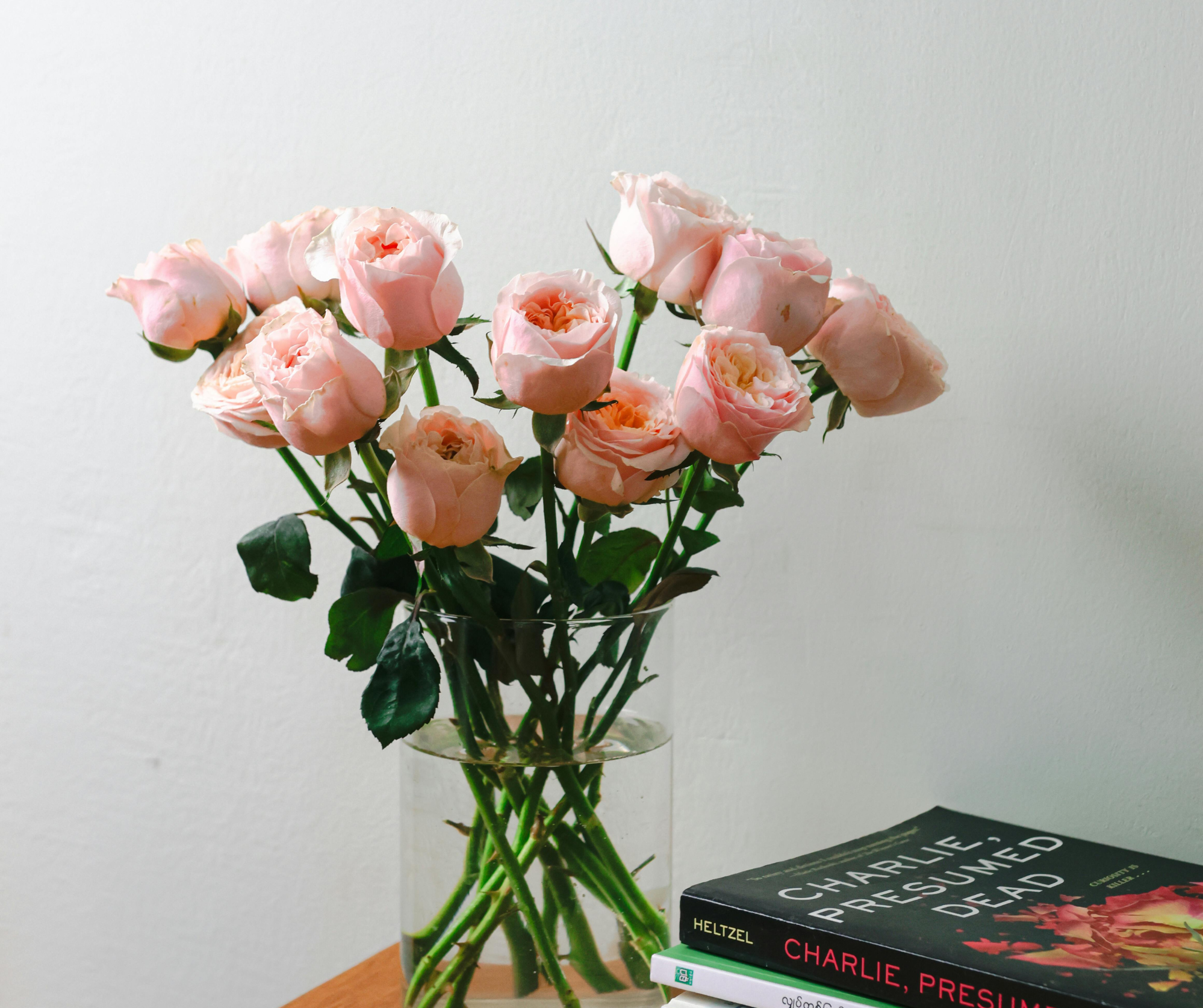 A bouquet of pink roses in a clear glass vase on a wooden surface, with a black and red book titled 'Charlie, Presumed Dead' nearby against a plain white background.